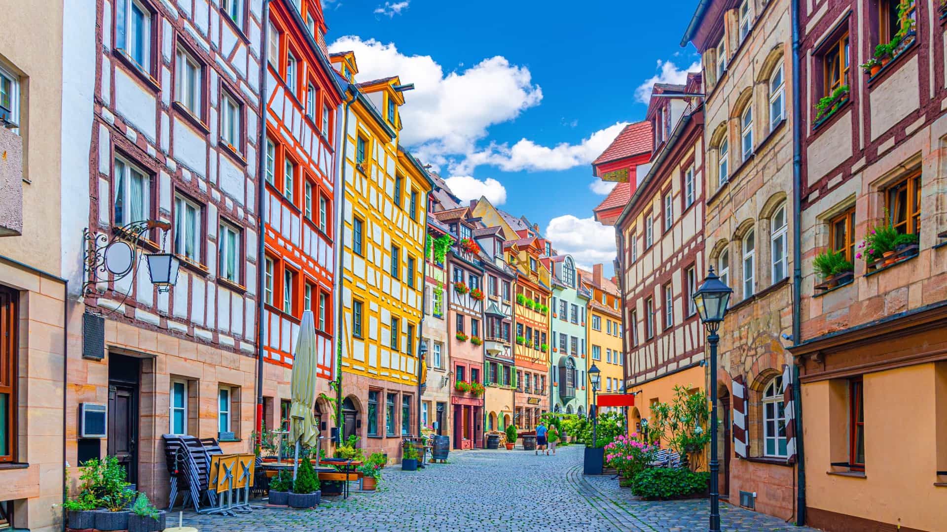 A view down the cobblestone street of Weißgerbergasse in the Old Town of Nuremberg, Germany. The narrow street is lined with a row of colorful, well preserved half timbered houses with red tiled roofs. Some of the buildings have shops on the ground floor, and the scene is set against a bright sky.