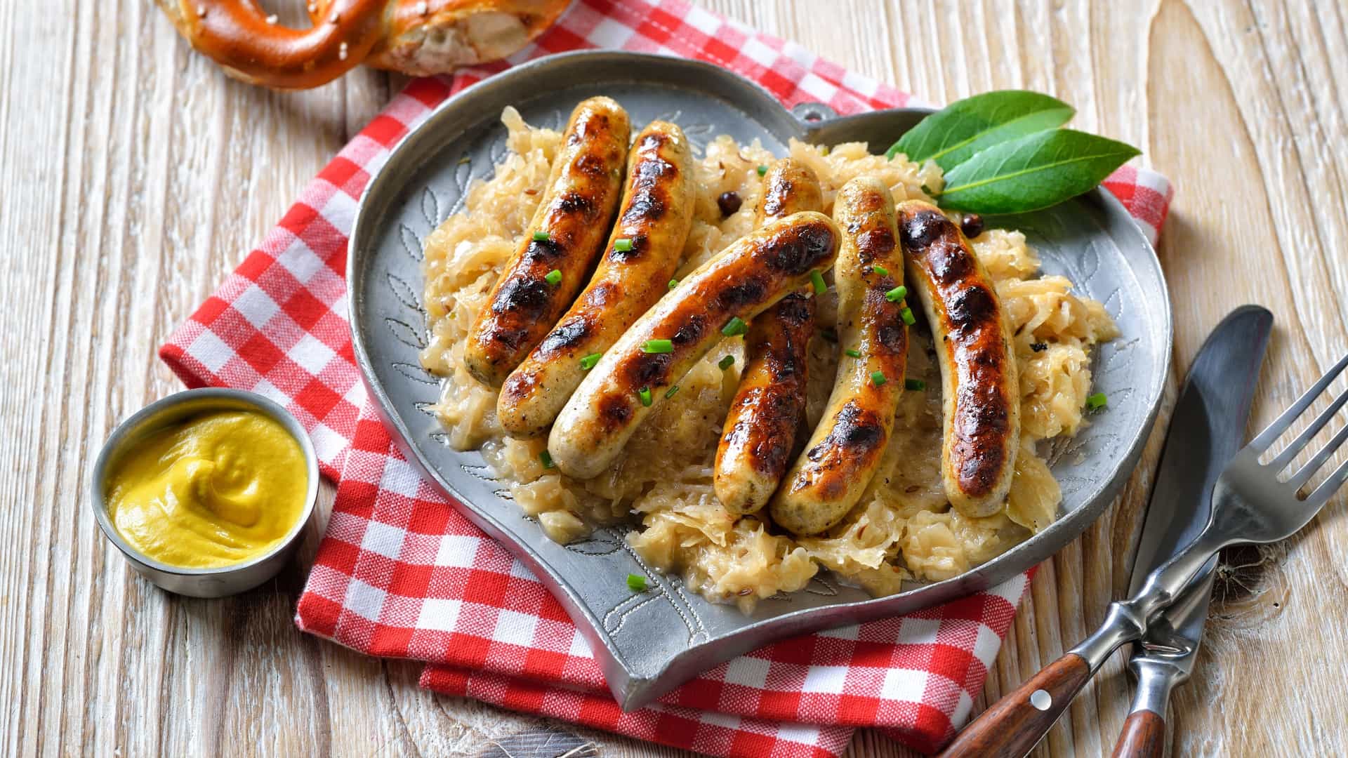 A photograph of six small, browned Nuremberg Rostbratwürste (fried sausages) neatly arranged on a mound of warm, tangy sauerkraut. The dish is presented on a simple white plate, ready to be served as a traditional German meal.