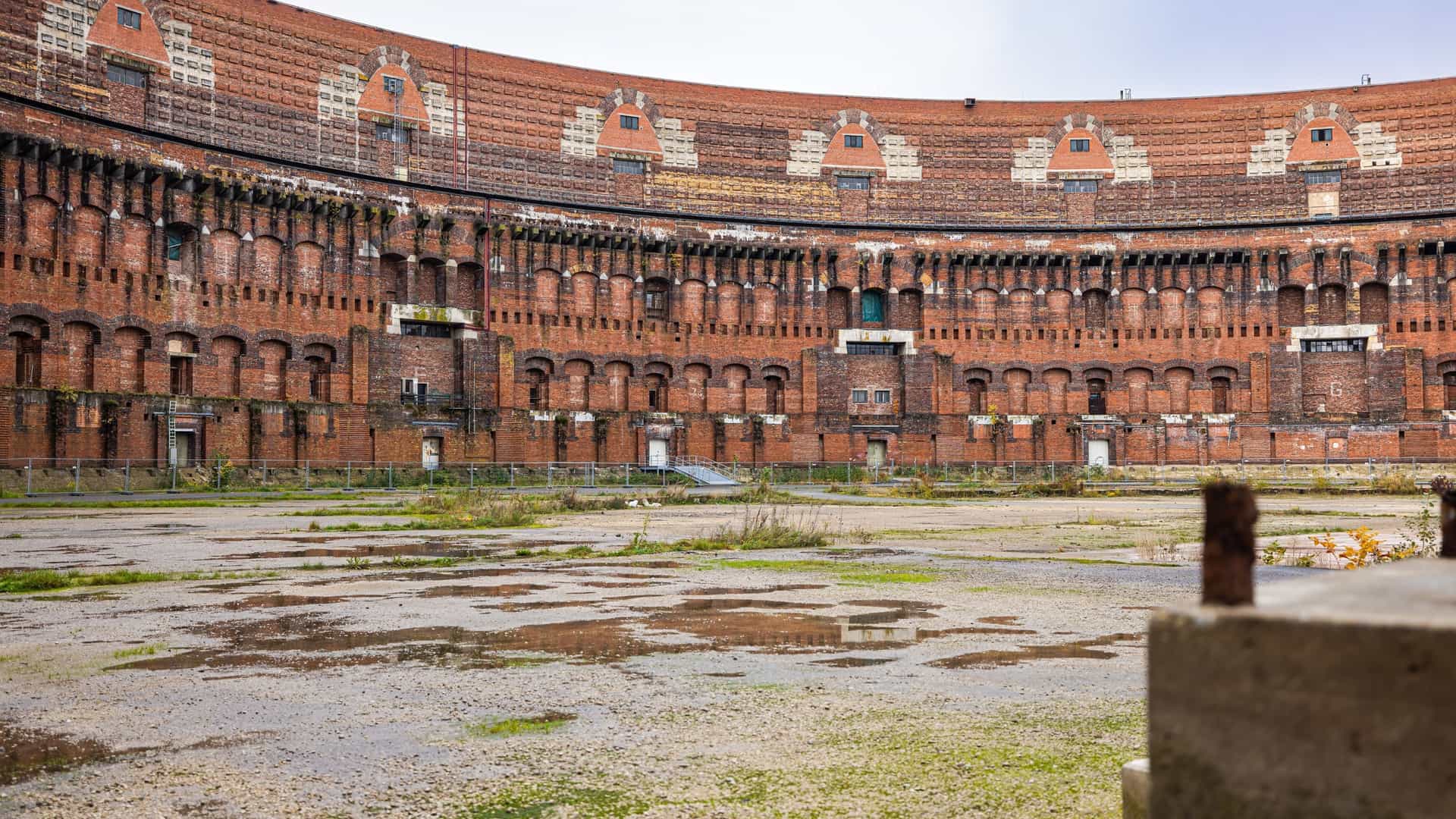 A large, unfinished, U shaped building known as the Congress Hall, located at the former Nazi Party Rally Grounds in Nuremberg, Germany. The building is a massive, monumental structure made of dark stone and is inspired by the Roman Colosseum. It stands next to a body of water under a clear sky. A modern, glass and steel addition, which houses the Documentation Center, is visible on the side of the historic building.
