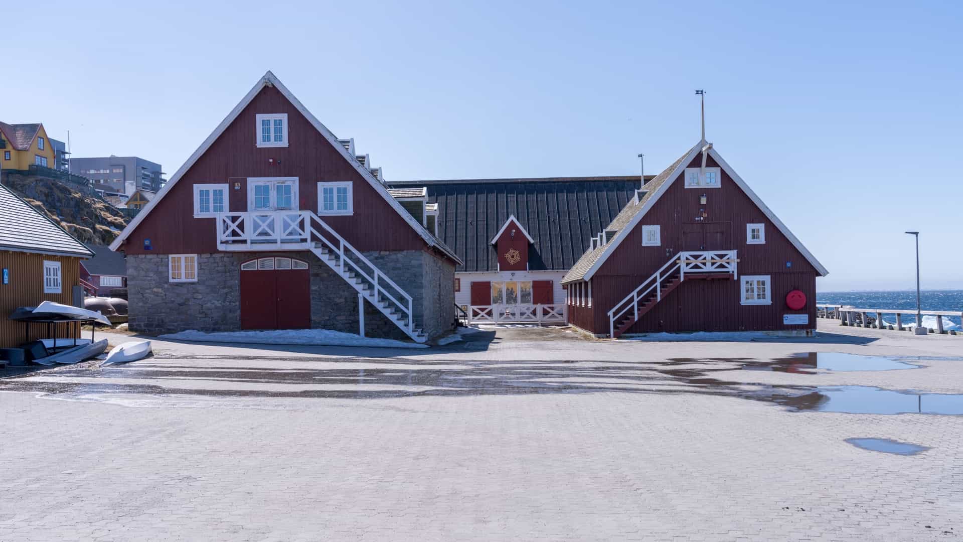 A view of the Greenland National Museum & Archives in Nuuk, Greenland. The museum is a collection of historic, red and white colonial style buildings situated at the edge of a fjord. The buildings have red roofs and are surrounded by a paved area and scattered patches of grass, with the calm water of the fjord in the foreground.