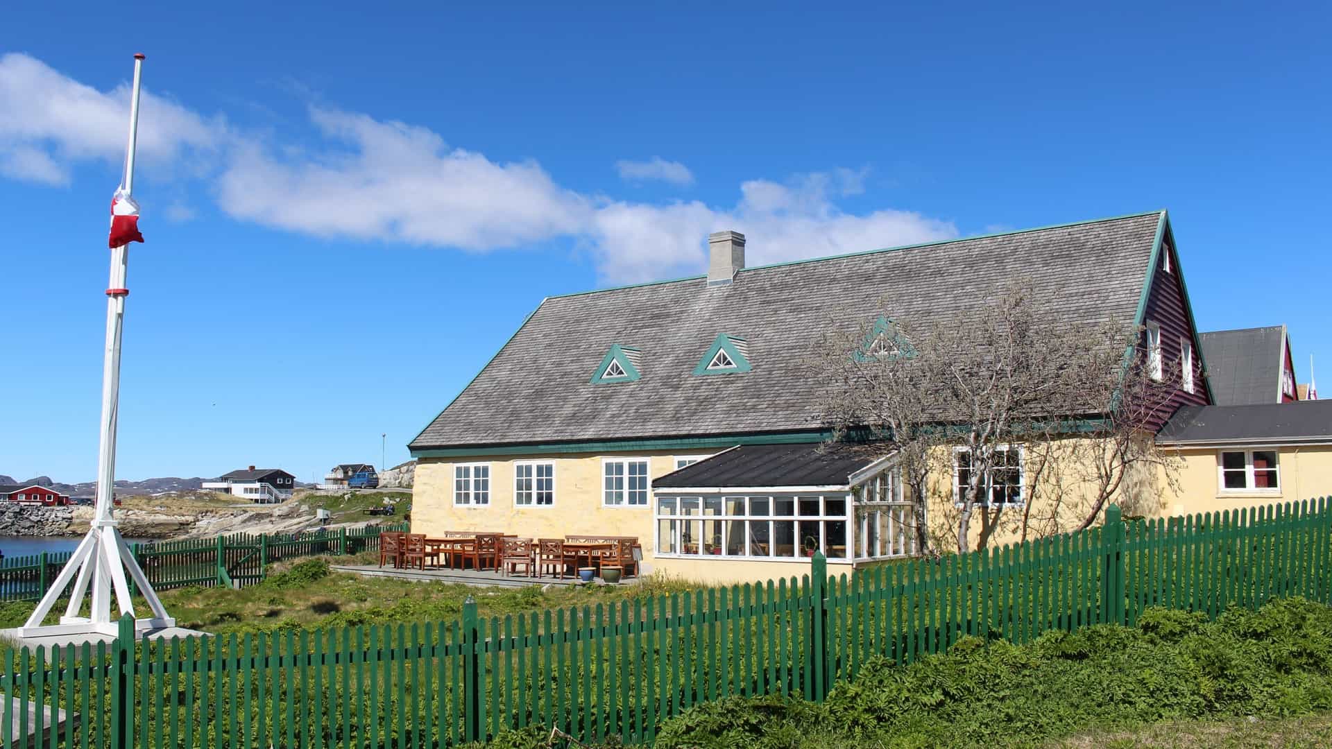 A sunny, wide-angle view of the historic Hans Egede House in Nuuk, Greenland, the country's oldest house. The large, two-story yellow building features a dark gray shingled roof, white-trimmed windows, and a glass-enclosed porch. In the foreground, a vibrant green picket fence surrounds a lawn, and a tall white flagpole with the Greenlandic flag stands to the left, set against a bright blue sky over the coast.