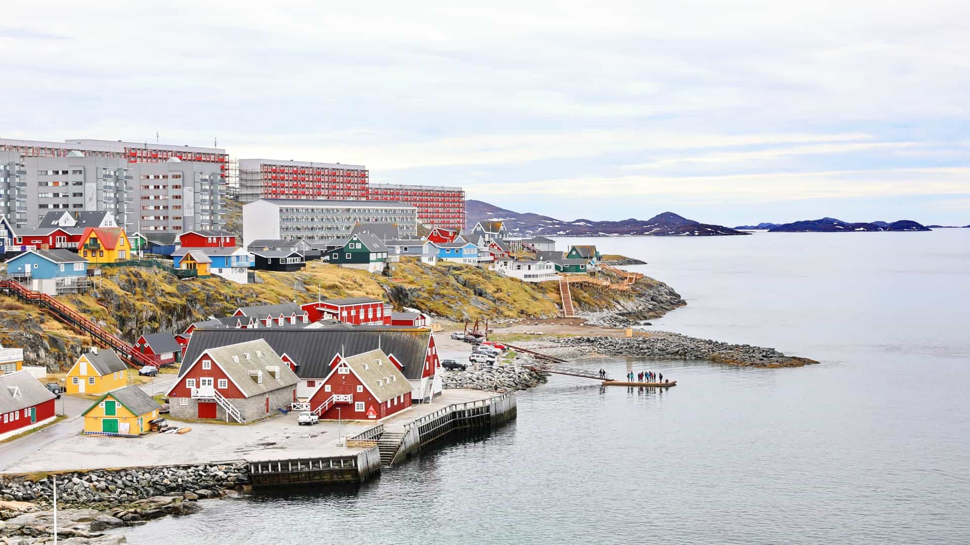 A scenic view of the old colonial harbor in Nuuk, Greenland. The waterfront is lined with a row of brightly colored buildings, including houses and other structures, with red, blue, and yellow facades. The buildings are located at the base of a rocky, dark colored hill, with snow capped mountains visible in the distance across a calm body of water.