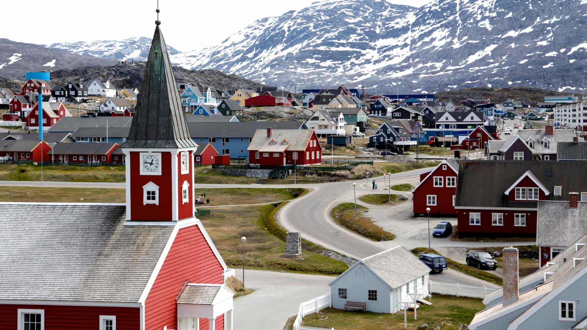 A view of the Nuuk Cathedral (Church of Our Saviour) in Nuuk, Greenland. The wooden, red painted church with a white spire stands prominently against a backdrop of a cloudy sky and mountains. The church is situated on a rocky outcrop, and its red color contrasts with the surrounding green landscape.