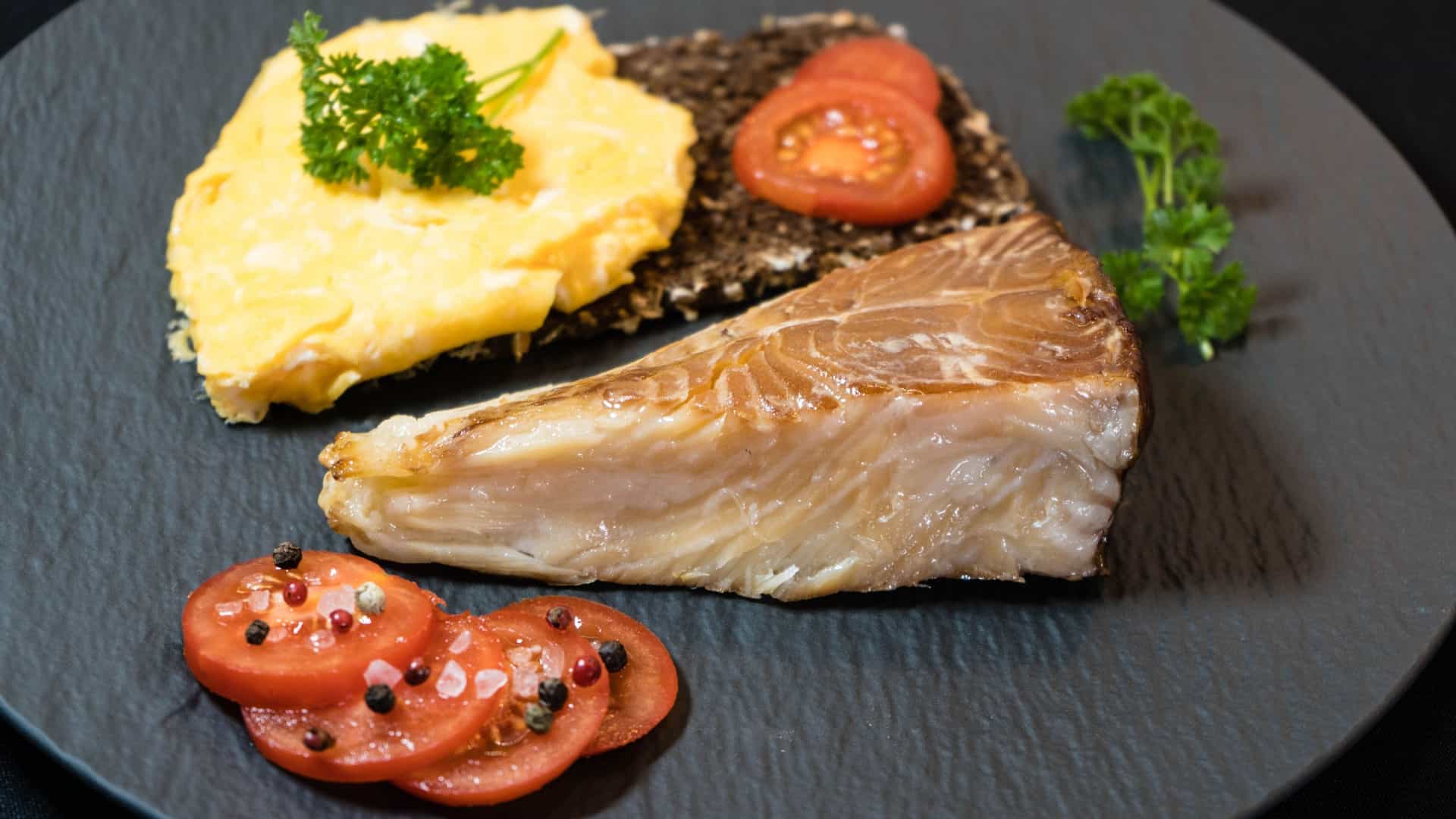 A photograph of a plate featuring a piece of white, flaky smoked Greenland halibut, served alongside a fresh tomato salad and a slice of bread. The dish highlights the local cuisine of Greenland, combining the prized regional fish with simple, fresh accompaniments.