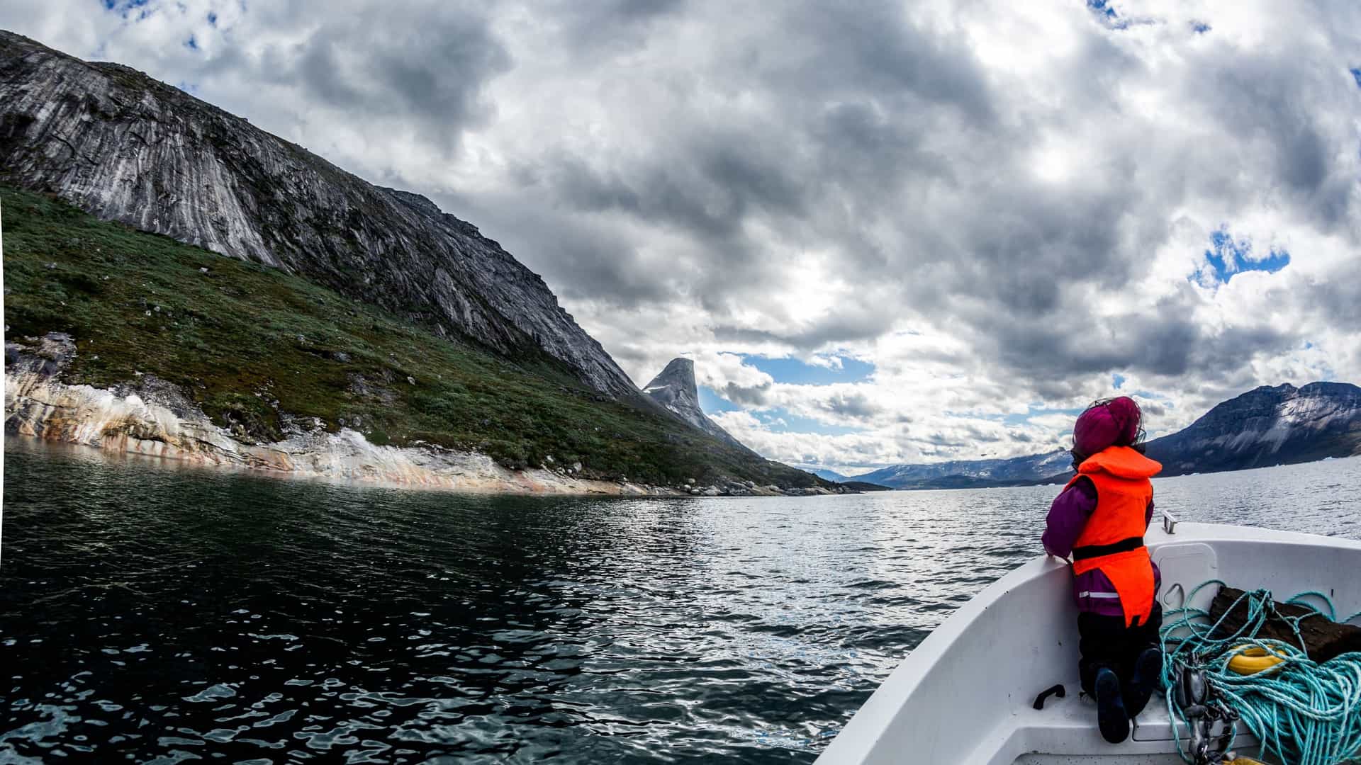 A scenic view of the Nuuk Fjord in Greenland, with a small boat on the calm water. The boat is cruising in a fjord surrounded by tall, steep mountains with rugged, rocky faces. The sky is partially cloudy, and the overall landscape conveys a sense of adventure and wilderness.