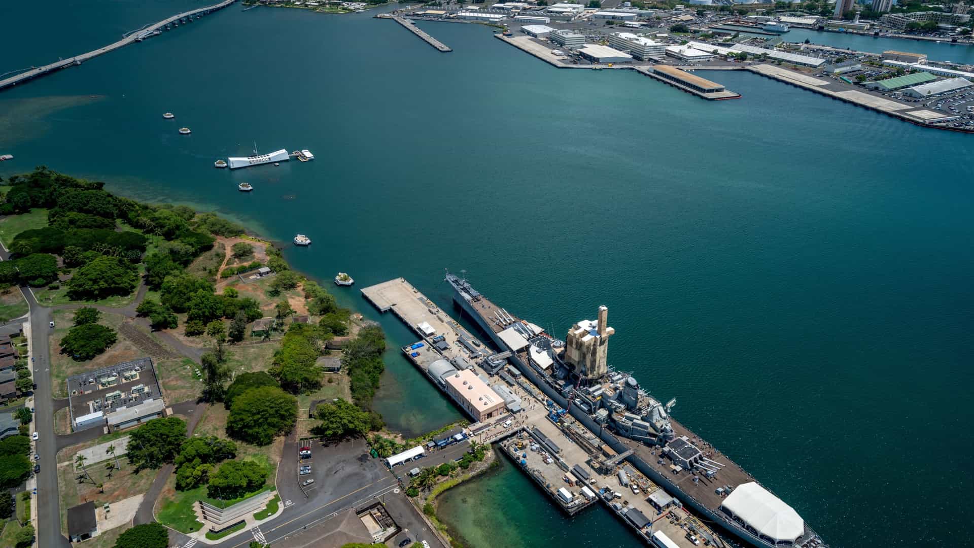 An aerial view of Pearl Harbor in Oahu, Hawaii, showing the expansive naval base. The image captures the blue water of the harbor with various ships and piers, surrounded by a peninsula and residential areas. In the distance, a highway and buildings of Honolulu are visible, set against the backdrop of the ocean.