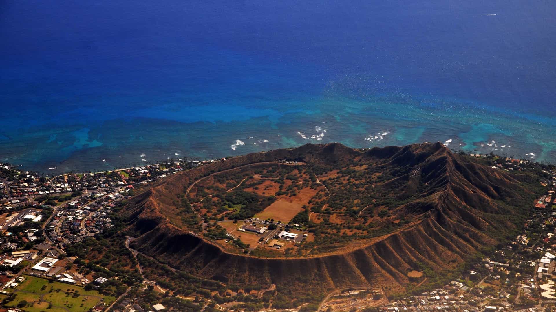 An aerial view of Diamond Head, the distinctive, extinct volcanic tuff cone that dominates the skyline of Oʻahu, Hawaiʻi. The image shows the horseshoe-shaped crater, covered in dry green brush, rising steeply from the coastline. The clear blue Pacific Ocean and a portion of Waikiki Beach and the city are visible in the background.