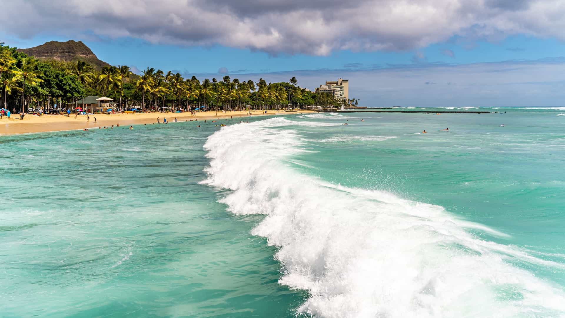 A high angle view of Waikiki Beach in Honolulu, Hawaii, showing a sandy shoreline filled with people relaxing on lounge chairs and swimming in the calm, turquoise water. Palm trees line the beach, and the background is filled with high rise hotels and resort buildings. The iconic Diamond Head volcano is visible in the distance under a clear sky.