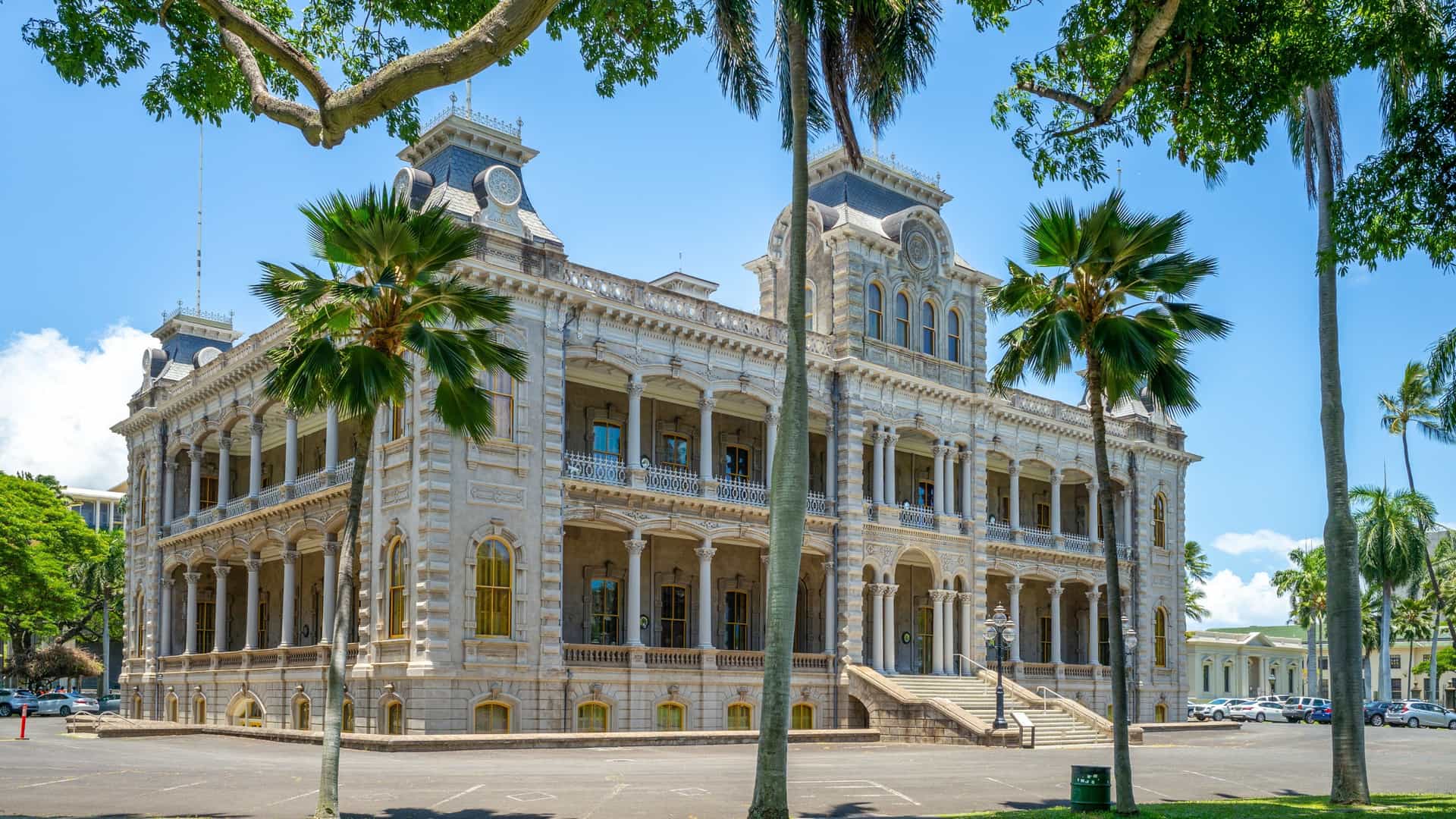 A view of Iolani Palace in Honolulu's Historic Downtown, Hawaii. The historic building, with its unique American Florentine architectural style, features four corner towers and a detailed exterior. It is surrounded by a manicured lawn with tropical trees and a path leading to the entrance. The palace is a symbol of Hawaiian royalty and a significant landmark.