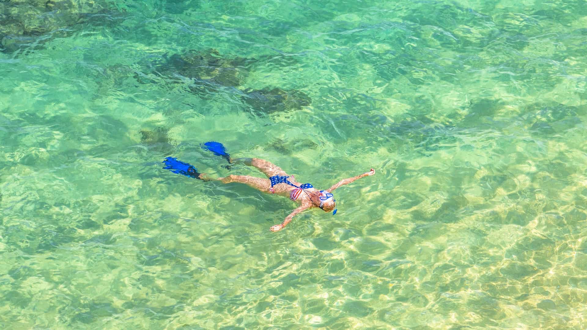 A high angle underwater view of a woman snorkeling over a vibrant coral reef in the clear, turquoise waters of Hanauma Bay Nature Preserve, Oahu, Hawaii. The woman, wearing a red, white, and blue bikini, is seen from above as she floats face down, exploring the diverse marine life and colorful corals below.