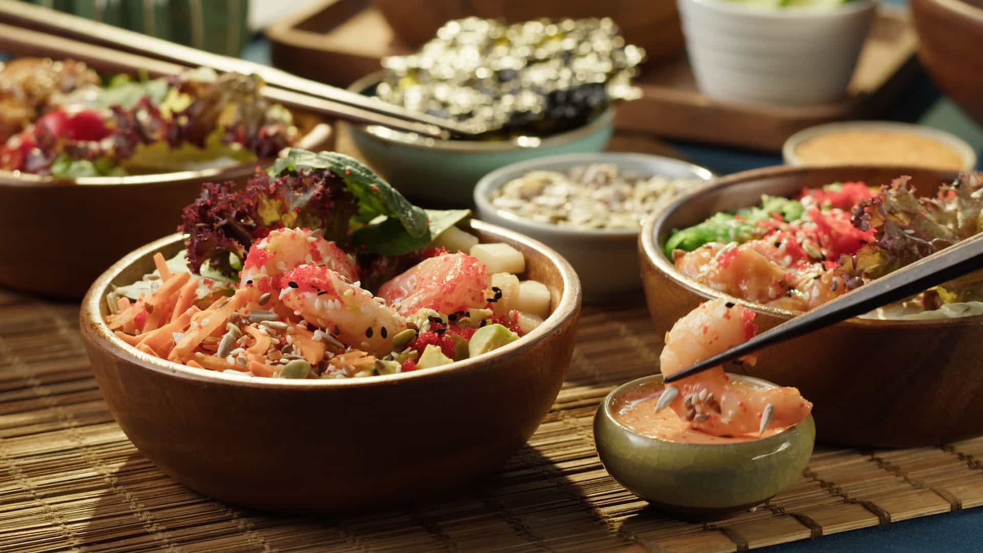 A close up, overhead shot of a person's hand sprinkling toasted sesame seeds over a freshly prepared poke bowl. The bowl contains a base of cooked rice topped with cubed, cooked fish, and is garnished with other fresh ingredients, such as chopped green onions or vegetables. The focus is on the action of adding the final garnish.