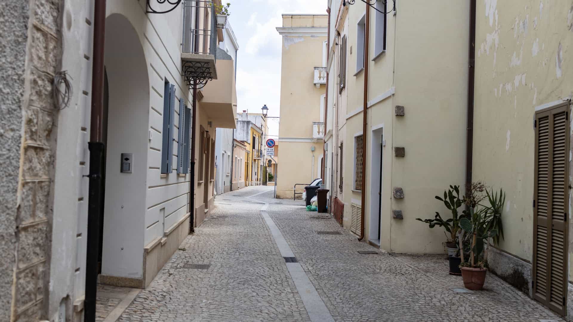 A photograph of a narrow, historic street in the Old Town of Olbia, Sardinia, Italy. The street is paved with rough stone or cobblestones and lined with traditional, light-colored buildings featuring balconies, wooden shutters, and perhaps small shops or cafes at ground level, suggesting a warm Mediterranean environment.