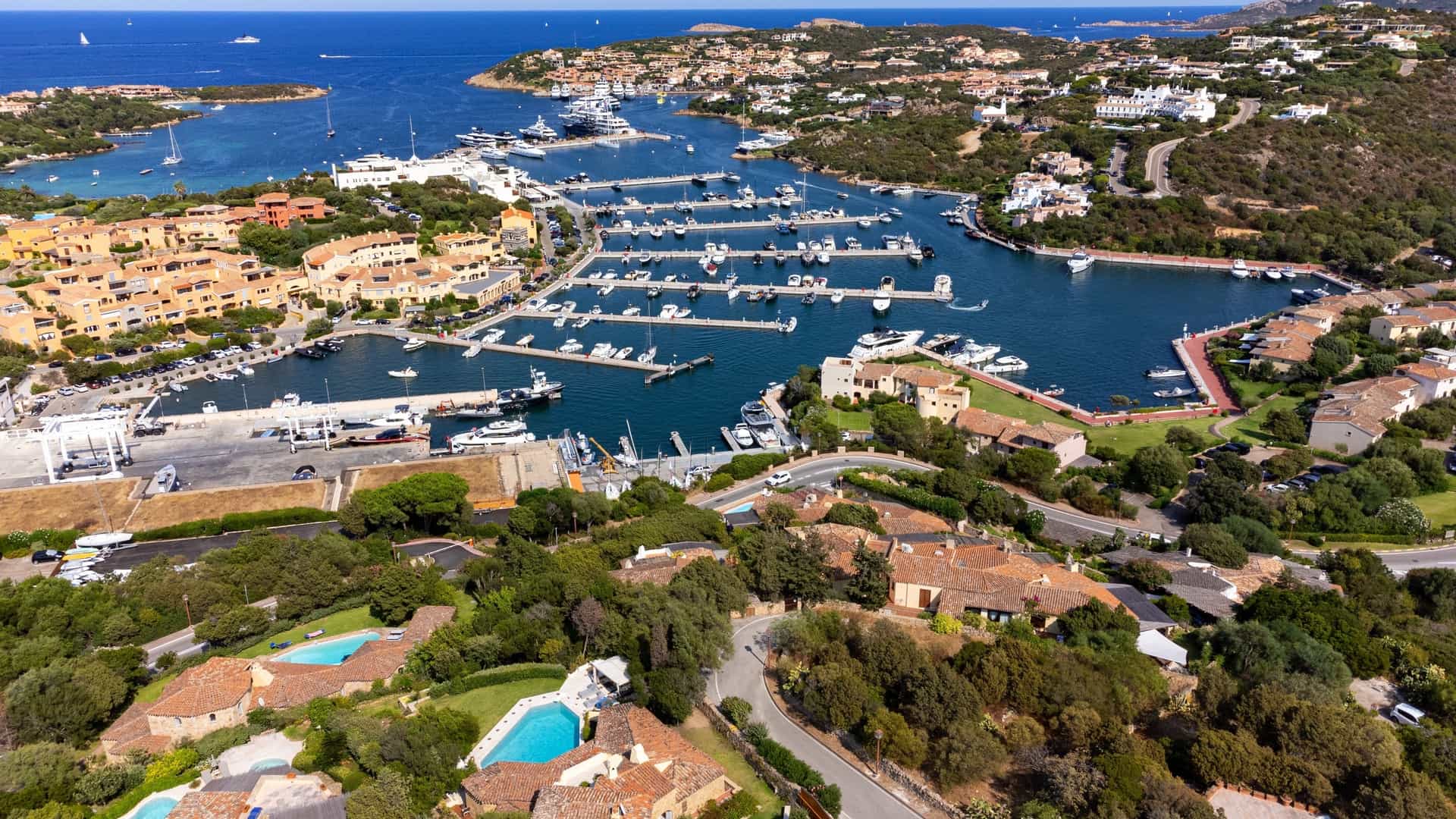 An elevated or aerial view of Porto Cervo, the main village on the Costa Smeralda in Sardinia, Italy. The image captures the town's distinct architecture of white and pastel-colored buildings with tiled roofs clustered around the deep blue harbor where numerous luxury yachts are moored. The rugged, emerald green coastline and clear Mediterranean Sea are prominently visible in the background.