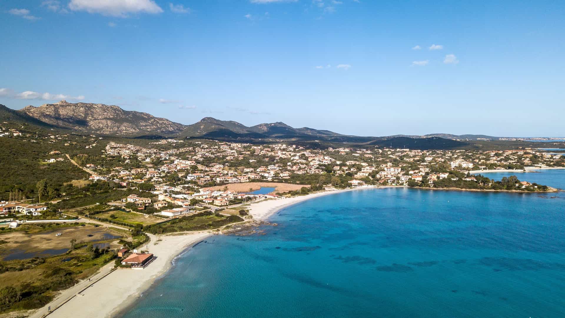 An aerial view of Pittulongu Beach near Olbia, Sardinia, Italy. The image shows the curved strip of fine, white sand contrasting sharply with the clear, shallow turquoise waters of the sea. The surrounding coastline is dotted with scattered buildings, indicating a popular coastal settlement.