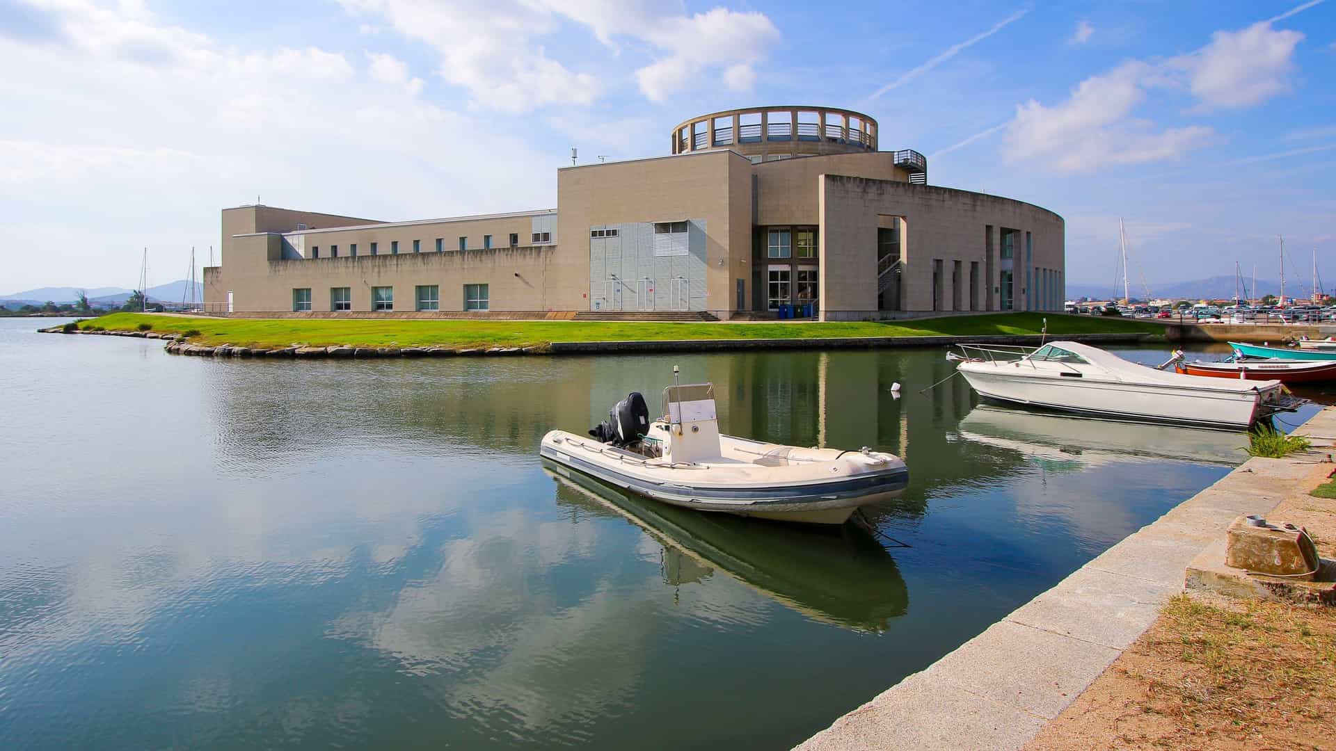 An exterior, architectural photograph of the Archaeological Museum of Olbia located on the island of Peddone in Sardinia, Italy. The museum is a modern building featuring a distinctive, angular facade with extensive use of glass and steel, designed to resemble a ship, reflecting Olbia's maritime history. The structure stands prominently near the water against a blue sky.