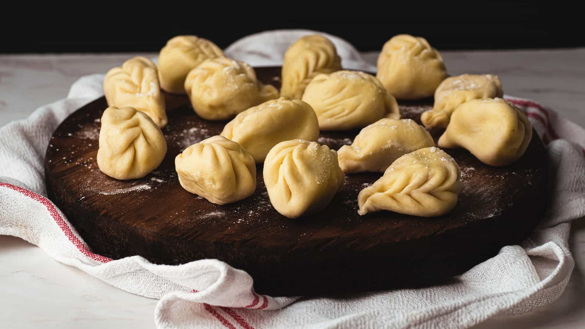 A close-up photograph of a plate of Culurgiones, the typical stuffed pasta of Sardinia, Italy. The pasta pieces are characterized by their distinctive, wheat-ear-shaped, crimped edges. They are likely filled with potato, mint, and cheese, and are shown plated as a main course, perhaps drizzled with tomato sauce and olive oil.