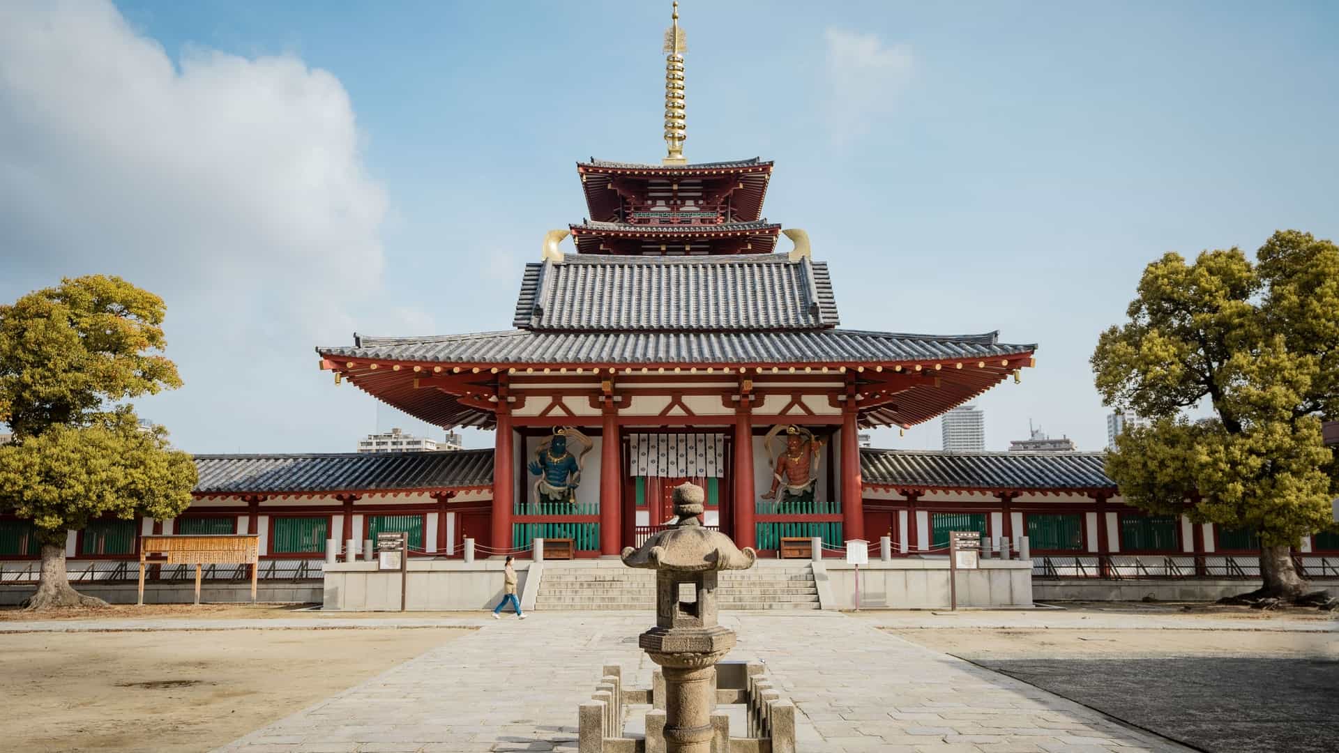 A view of Shitennoji Temple in Osaka, Japan. The central focus is the temple's iconic five story pagoda, a tall, tiered structure with a vibrant vermilion and white exterior and green roofing. The pagoda is a prominent feature of the temple's grounds, surrounded by other traditional Japanese buildings and pathways. The image captures the historical and cultural significance of one of Japan's oldest temples.