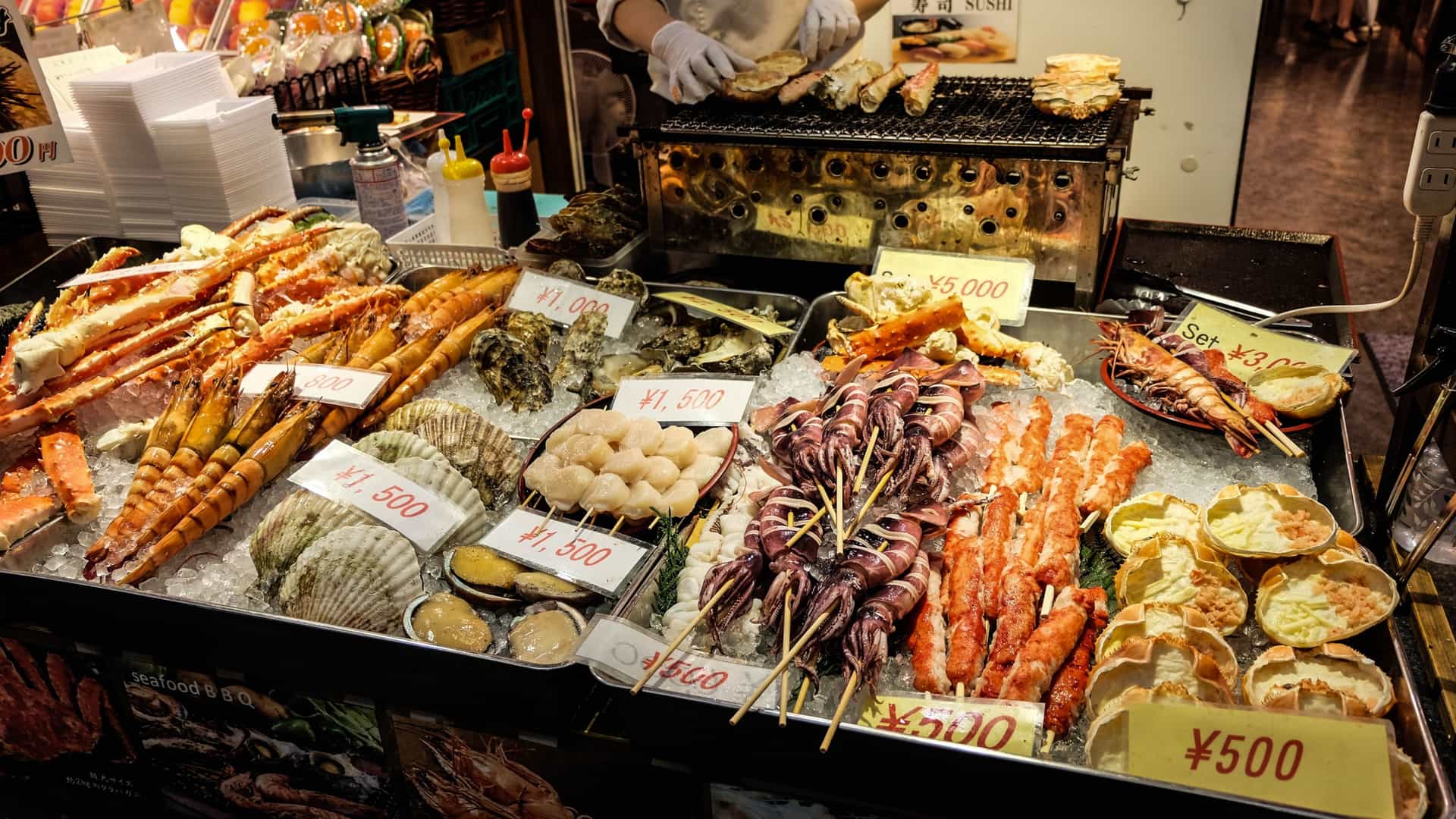A close up, low angle shot of a vendor's stall at Kuromon Ichiba Market in Osaka, Japan, showcasing a colorful variety of fresh seafood. The image captures a detailed array of raw shellfish and fish on ice, with some items displayed on skewers, ready to be grilled and eaten. The vibrant colors of the seafood, including a large, bright orange crab, stand out against the blue of the ice and the background of the market stall.
