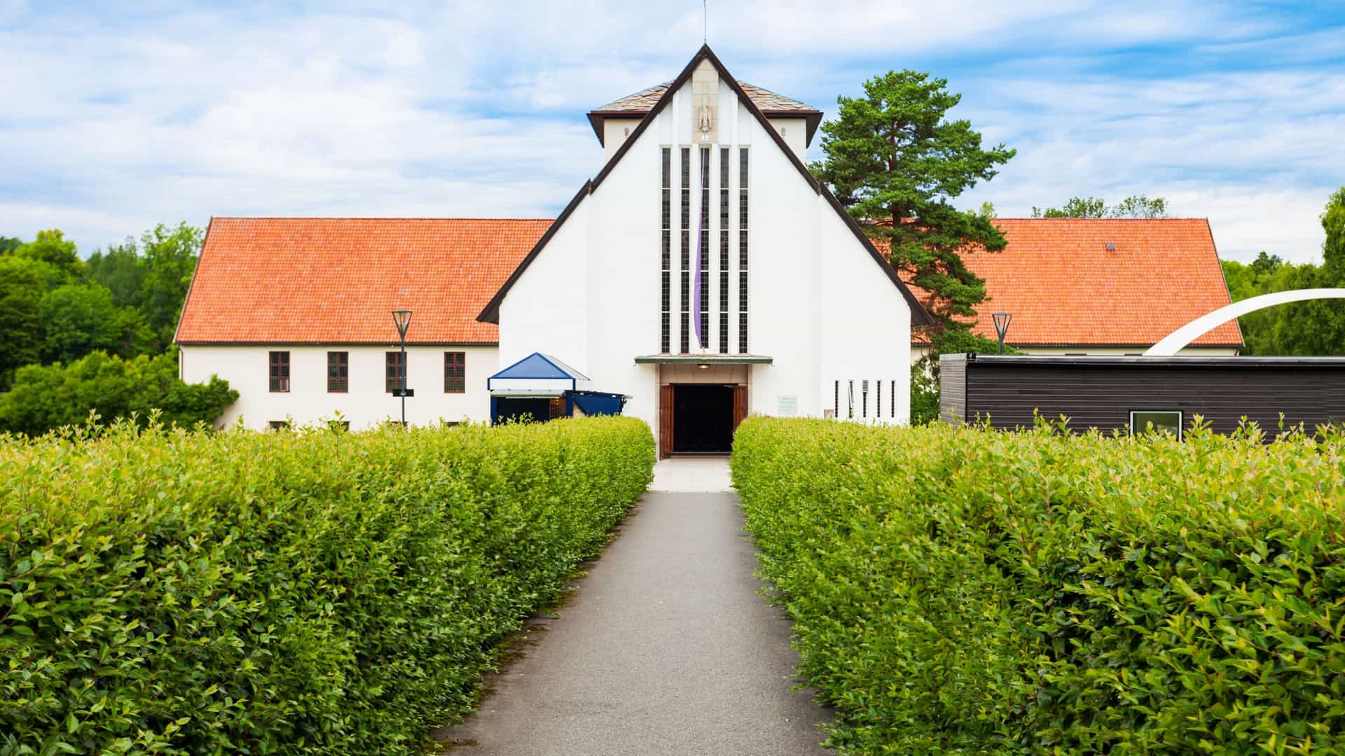 An exterior view of the Viking Ship Museum (Vikingskipshuset) located on Bygdøy peninsula in Oslo, Norway. The museum is a distinctive, low, white, cruciform concrete building with high central halls, built specifically to house and display the perfectly preserved Viking ships, visible through the large arched windows.