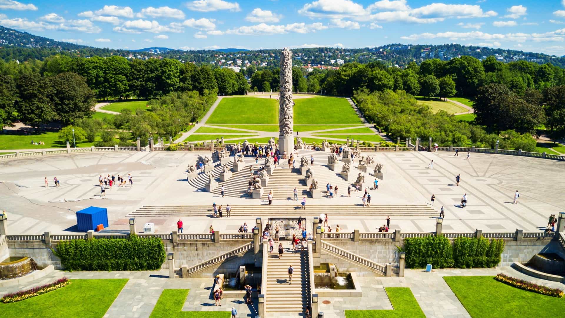 A serene, wide angle shot of a landscape in Vigeland Sculpture Park, Oslo, Norway. In the foreground, there are several large, life sized bronze and granite statues of human figures, some of which are intertwined with trees or other natural elements. The sculptures are positioned on a green lawn, and in the distance, a large, ornate fountain and the Monolith sculpture can be seen, surrounded by other figures. The sky is a clear blue, and the overall impression is one of art integrated with nature.