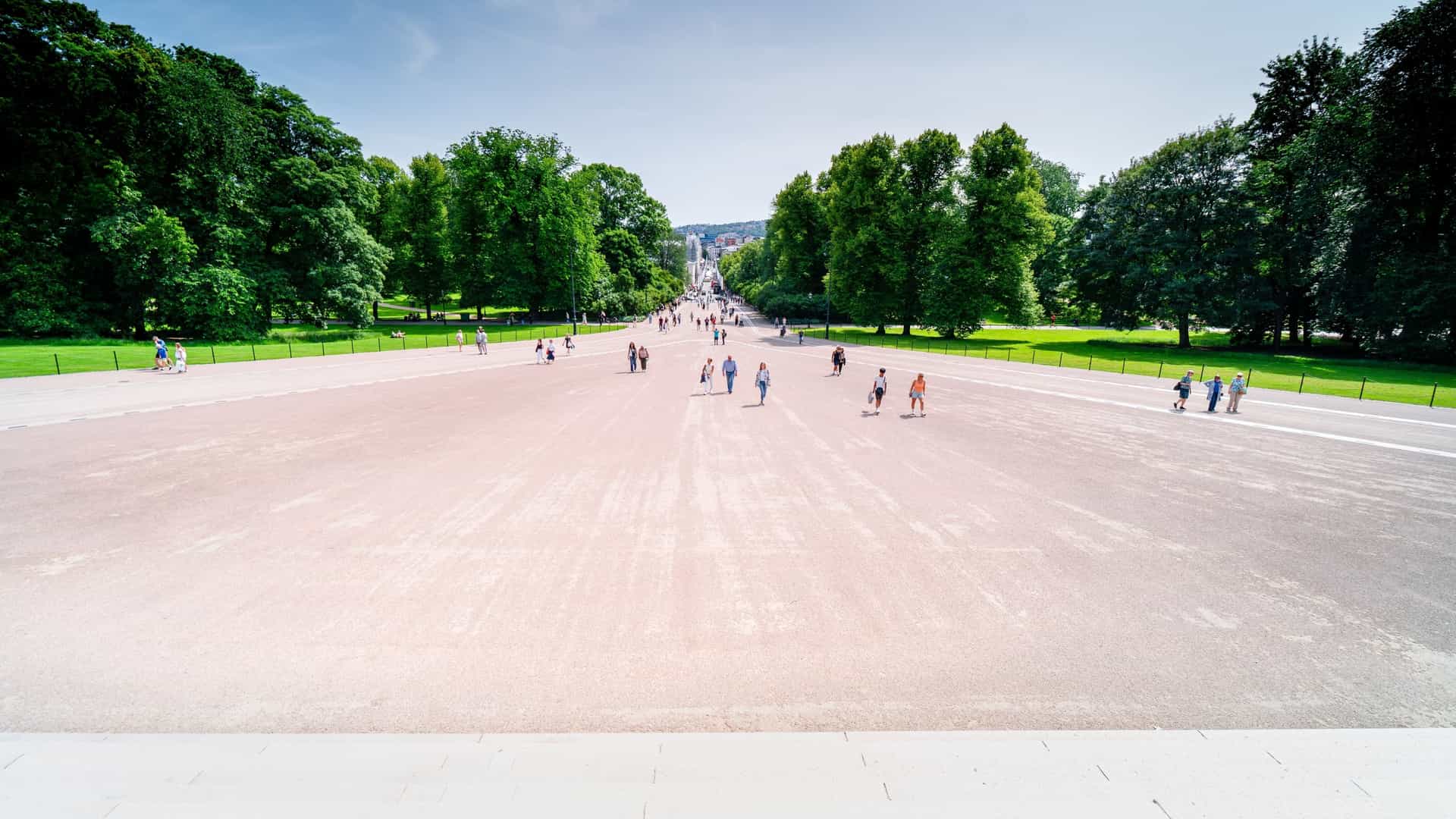 A sunny, eye level street view of Karl Johans gate, the main pedestrian thoroughfare in Oslo, Norway. The street is bustling with people walking and shopping. Historic European buildings with decorative facades line both sides of the street. Some people are seated at outdoor cafes, and a few trees are visible along the paved walkway, creating a lively urban scene.