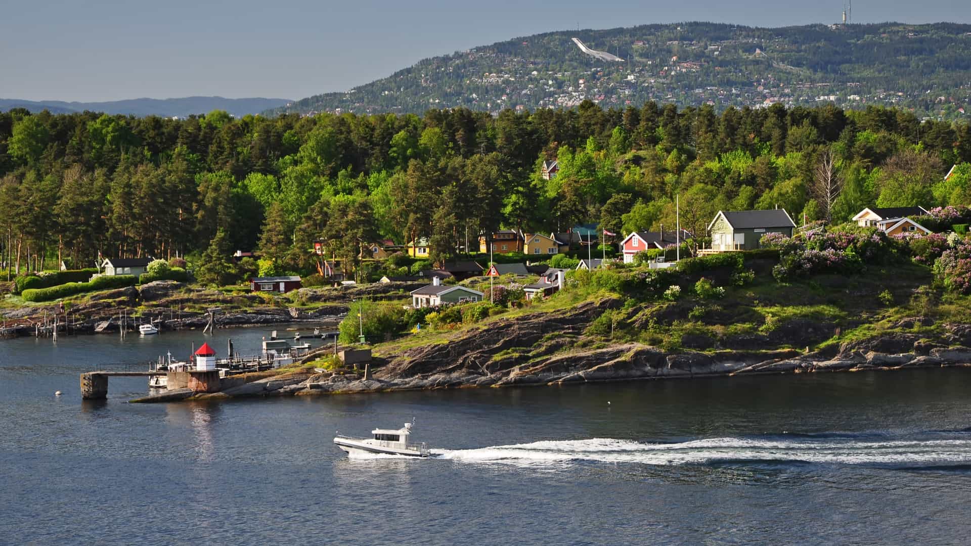 A scenic view of the Oslofjord in Norway. The foreground is a tranquil bay with calm, dark blue water. On the left, several traditional Nordic houses, painted in bright red and yellow, are built directly on a rocky outcrop. The houses have white trim and pitched roofs. The surrounding area consists of rugged, forested rock formations, with a clear sky and scattered clouds in the background.