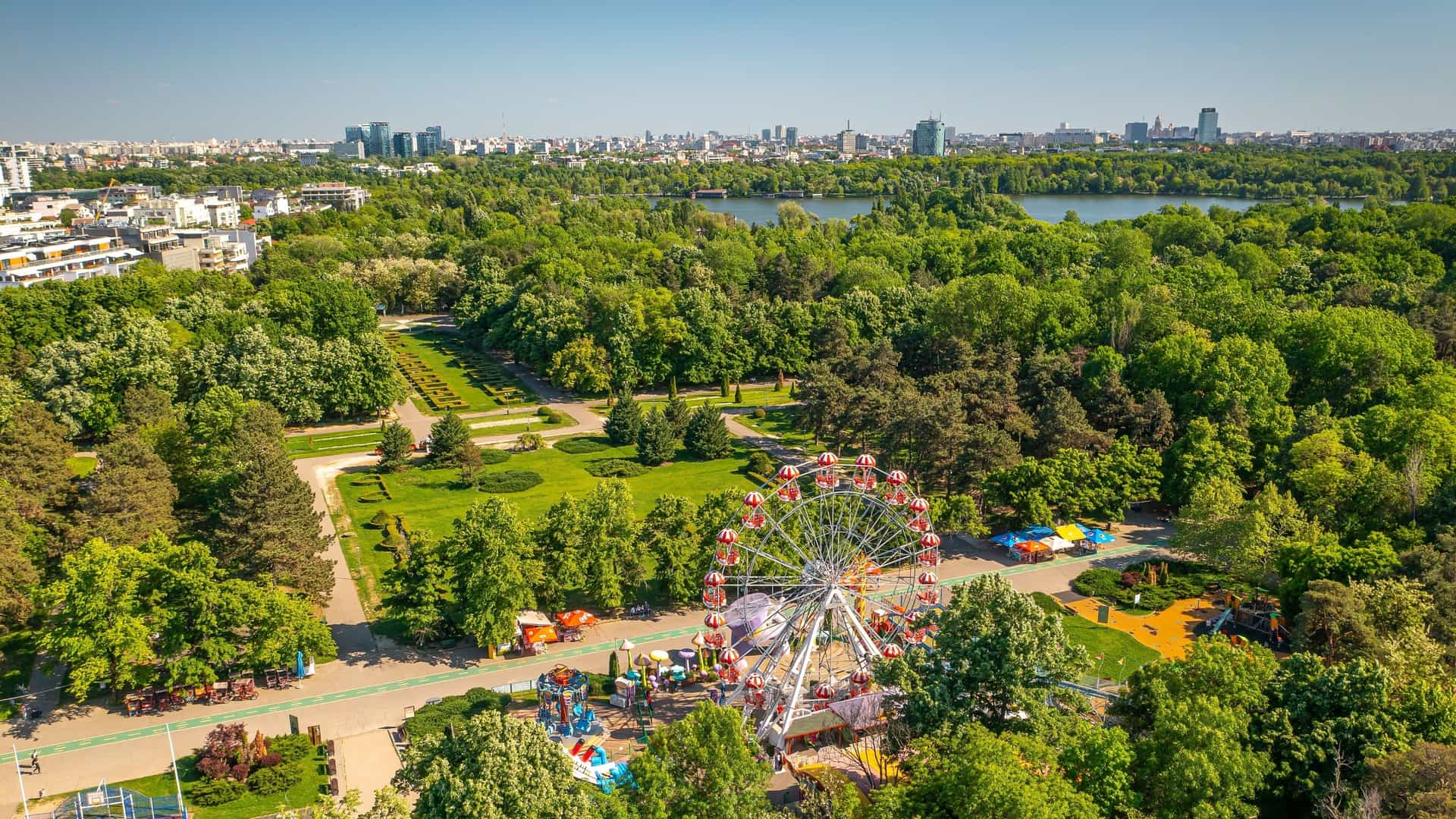 An aerial view of Herastrau Park in Bucharest, Romania, showcasing the large lake at its center, surrounded by lush green trees. In the background, the modern cityscape of northern Bucharest is visible, featuring various office buildings and high rise structures. The photo was taken on a sunny day.