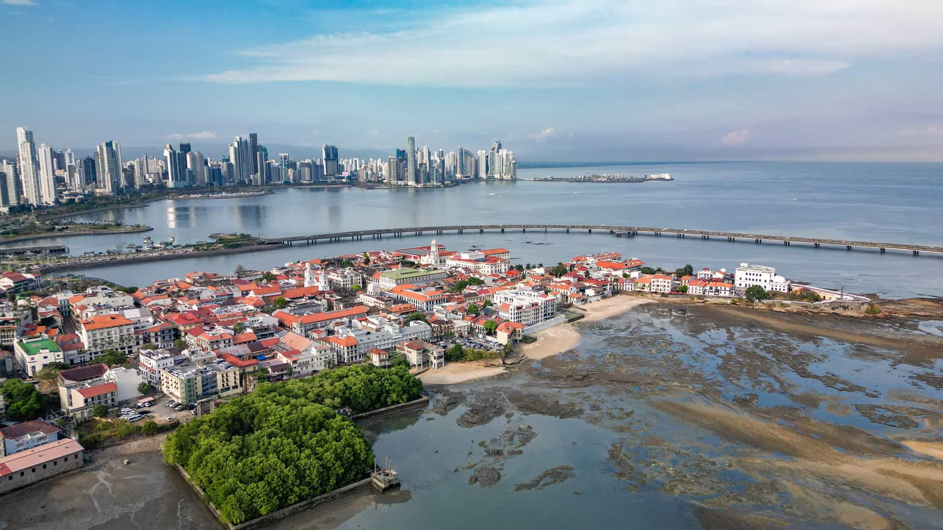 A panoramic view of the Casco Viejo (Old Quarter) of Panama City. In the foreground, the colorful, historic colonial buildings of Casco Viejo are visible, with their distinctive red tile roofs and traditional architecture. In the background, the modern skyline of Panama City rises dramatically, featuring numerous skyscrapers and high rise buildings along the coast. The image captures the striking contrast between the city's historic and contemporary districts.