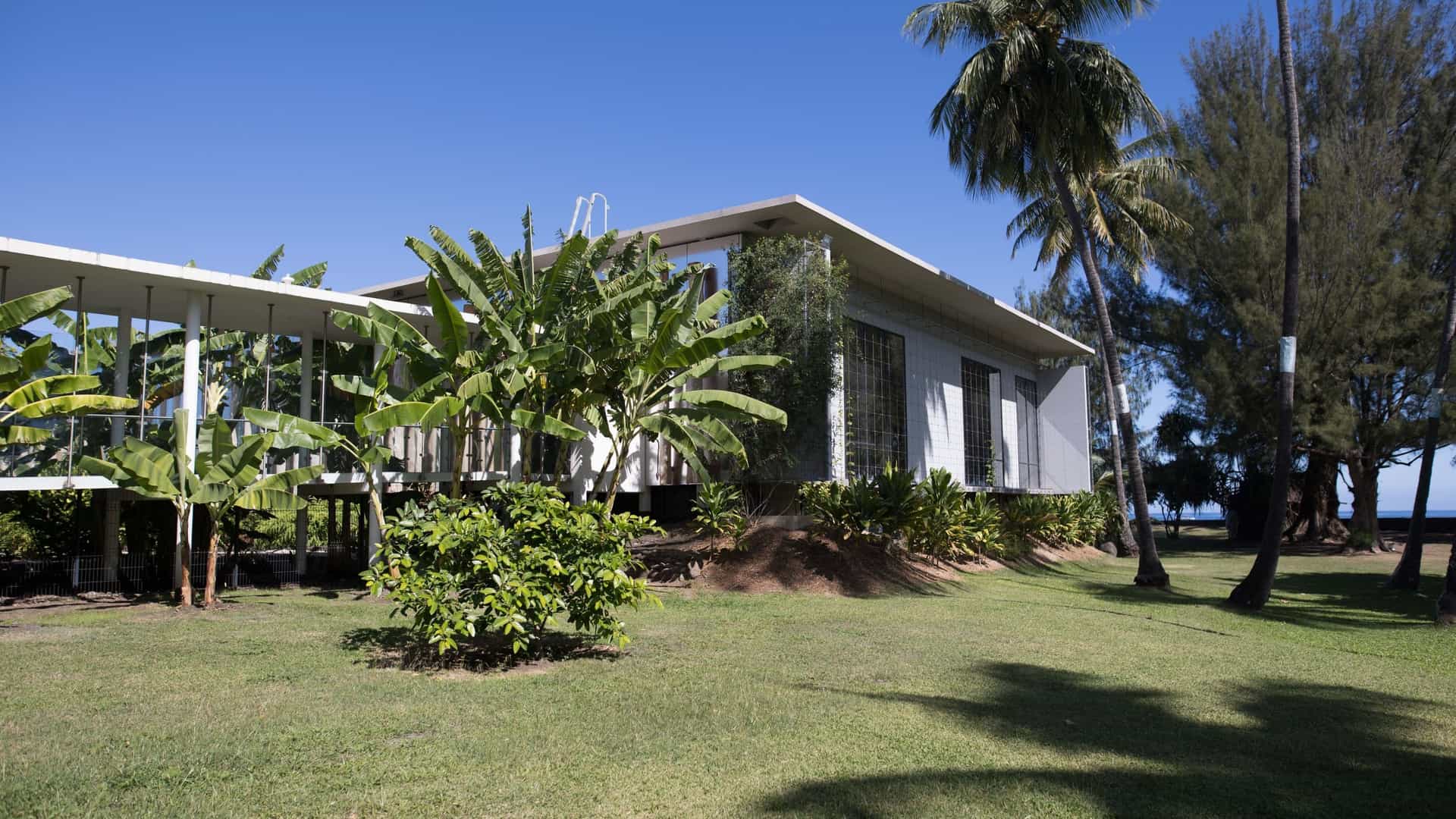 A view of the Te Fare Iamanaha Museum in Papeete, Tahiti, also known as the Museum of Tahiti and the Islands. The building features a modern, rectangular design with a large, sloping roof and expansive windows, set amid a lush green landscape. The museum is dedicated to Polynesian culture and history, and the surrounding area includes gardens and a view of the water, reflecting the island environment.