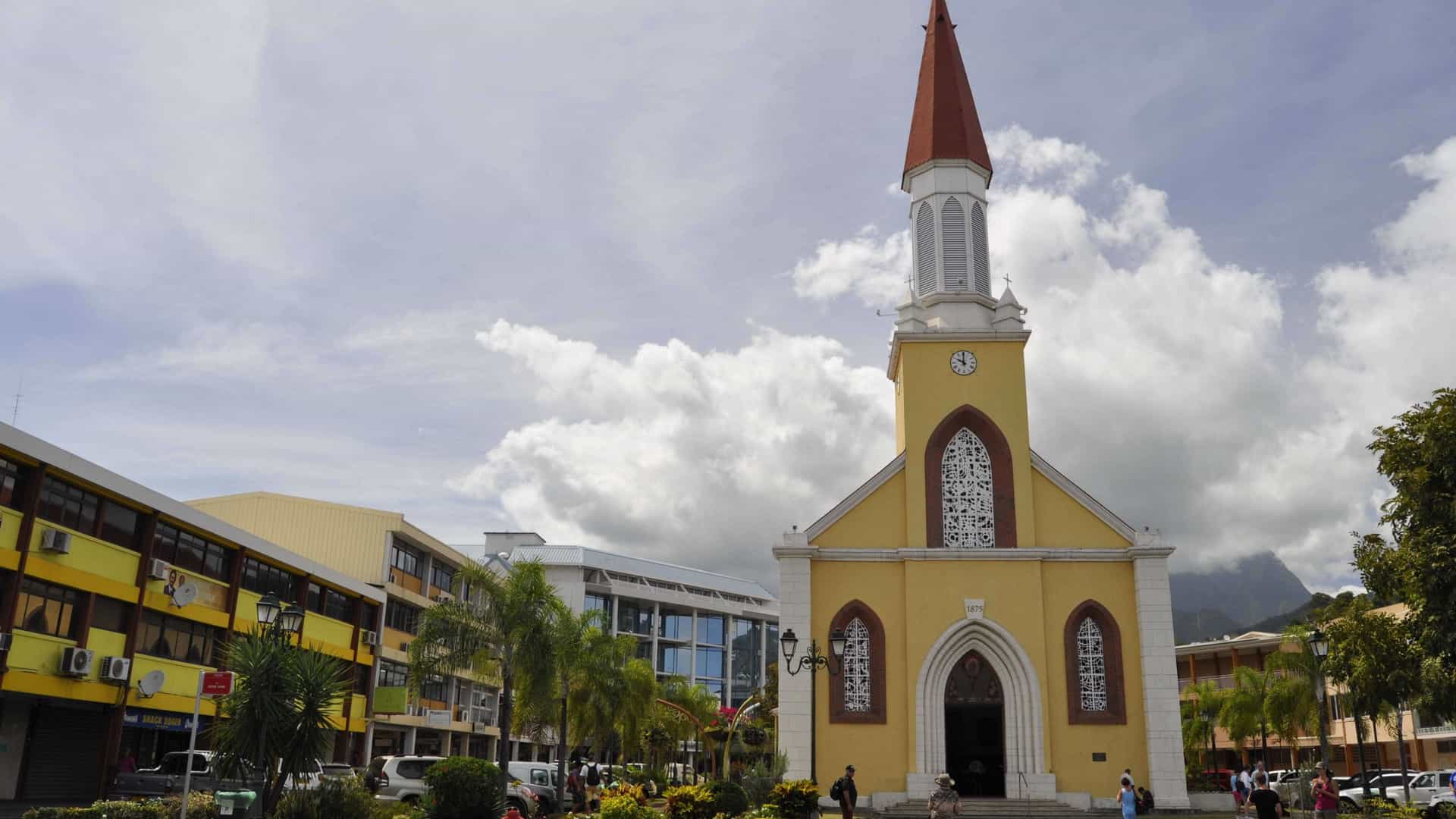 The exterior of the Notre Dame Cathedral in Papeete, Tahiti, a Catholic church with a grey facade and a prominent red tiled spire, standing against a bright blue sky. The photo shows the front of the church, with the entrance and a small courtyard in the foreground.