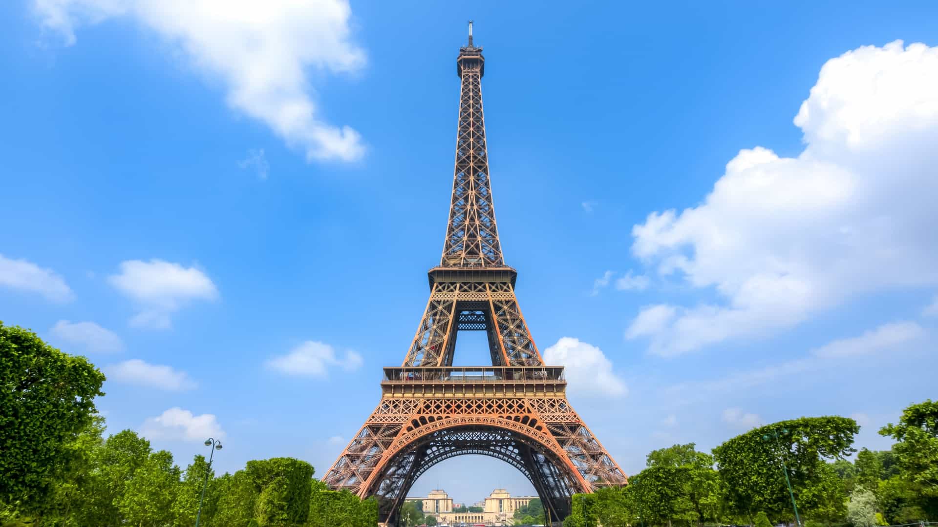 A scenic view of the Eiffel Tower from the Champ de Mars in Paris, France. The base of the wrought iron lattice tower is at the end of a vast, manicured green lawn, with a walking path and trees visible on the left side. The sky above is overcast.