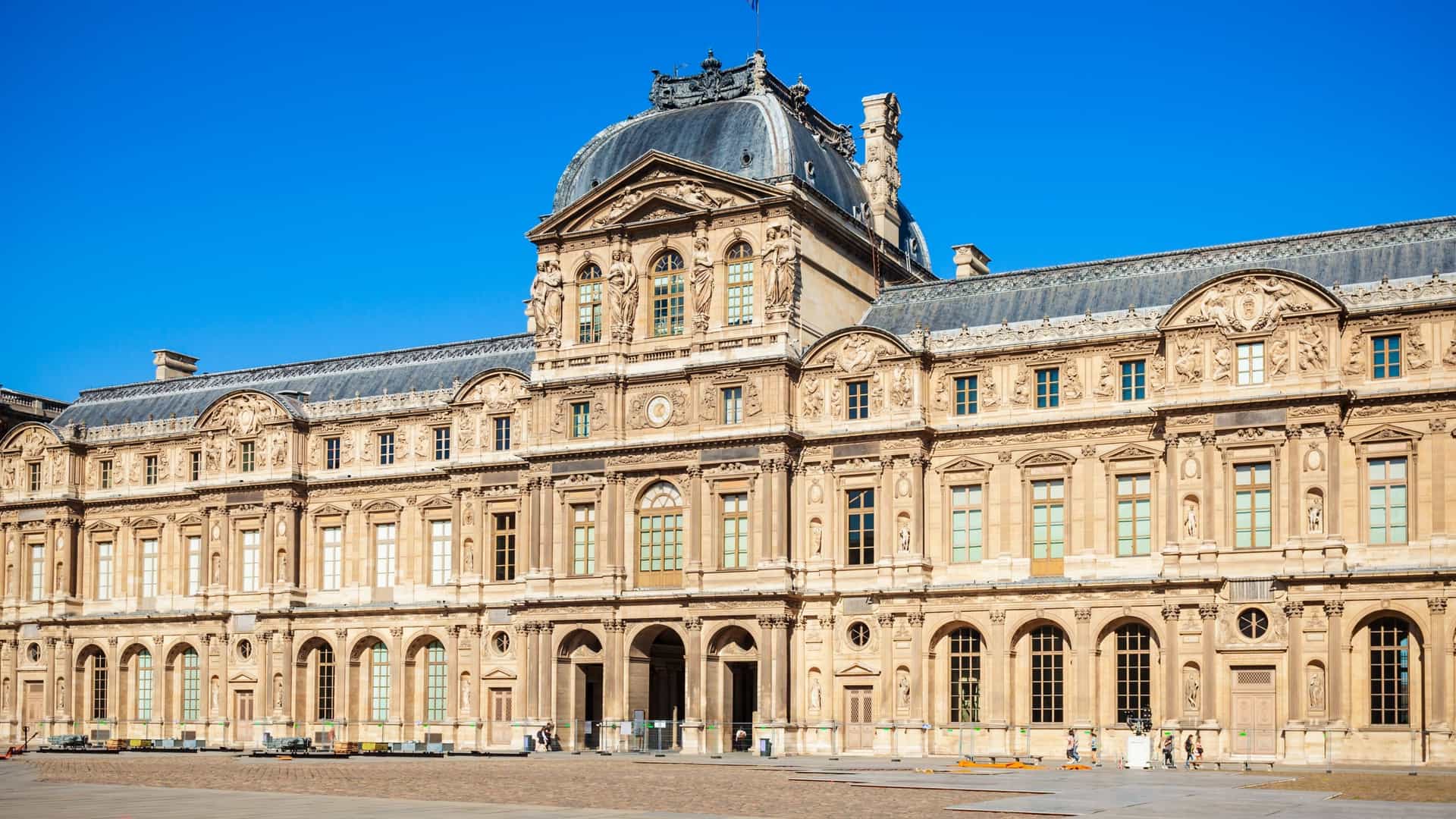 A view of the main courtyard of the Louvre Museum in Paris, featuring the iconic glass and steel pyramid designed by I.M. Pei. Fountains are visible at the base of the pyramid, with the classical architecture of the historic palace complex surrounding the courtyard under a bright sky.