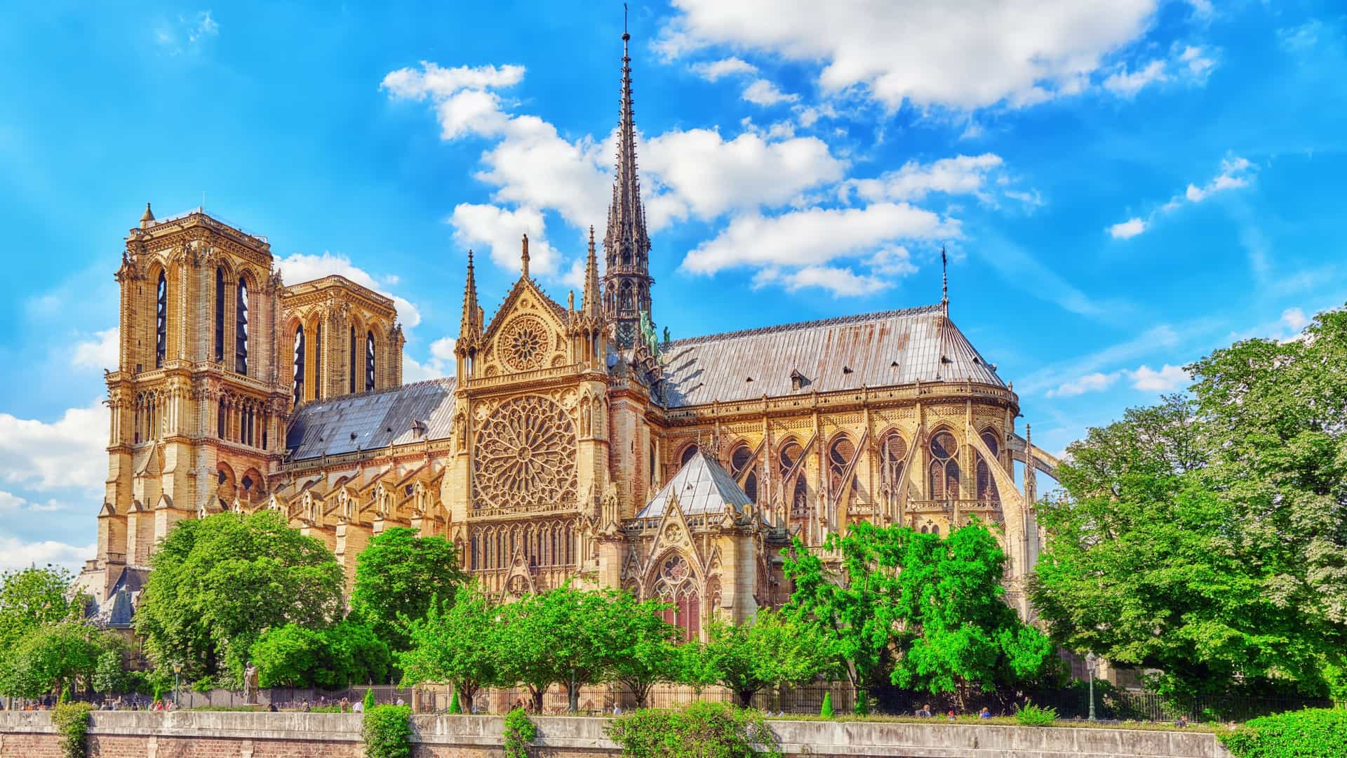 A view of Notre Dame de Paris Cathedral from the Seine River, showcasing its intricate Gothic facade, including the twin towers and a prominent rose window. The cathedral, a masterpiece of French Gothic architecture, stands majestically on the Île de la Cité.