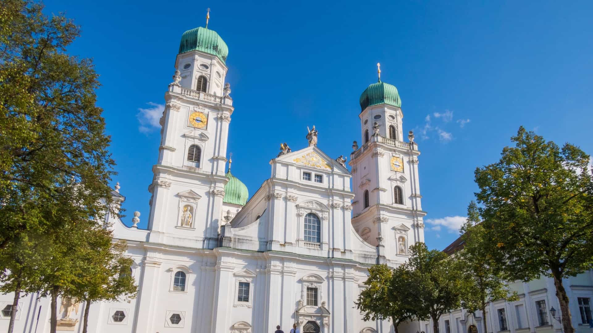 St. Stephen's Cathedral, a baroque church with white towers and blue green domes, sits on a hill in the old town of Passau, Germany. The historic city, known for its location at the confluence of three rivers, is visible in the foreground.