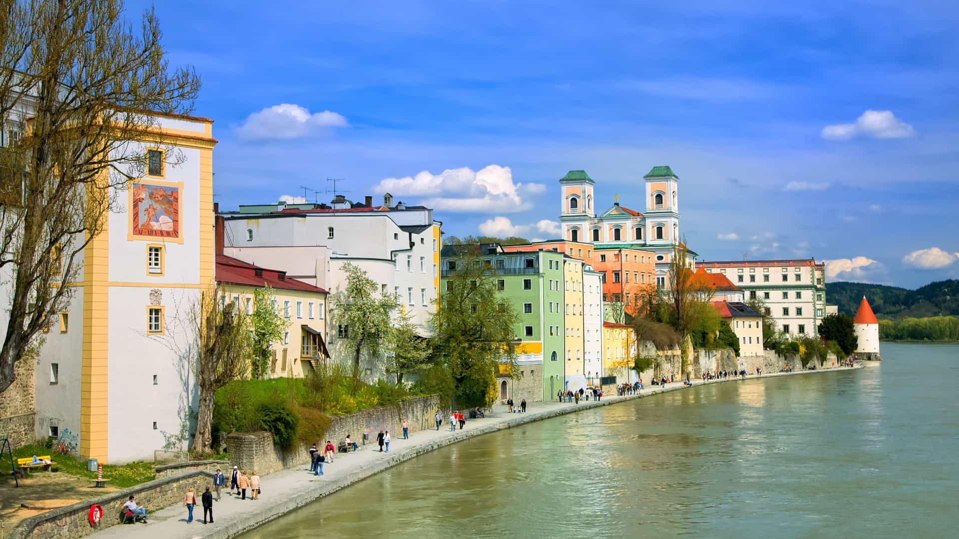 A wide, paved Danube River promenade lined with evenly spaced, trimmed trees and streetlights. Several people are seen enjoying a leisurely stroll along the path, with a view across the wide river to a dense and historic European city skyline, featuring prominent domes, classical buildings, and a mountainous backdrop.