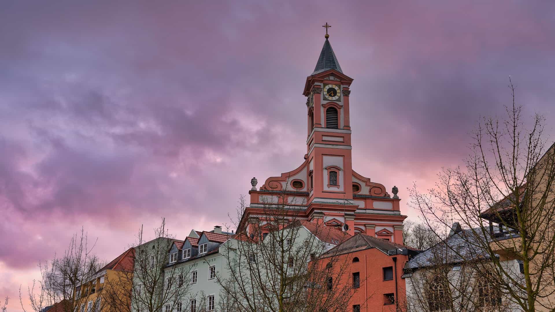 The Baroque exterior of the Parish Church of St. Paul in Passau, Germany. The church features a three-story tower with a copper-green spire and a facade painted in contrasting pastel tones of cream and pink. It is situated on a stone platform, with a covered flight of steps leading up to the entrance, and is surrounded by the narrow, historic buildings of Passau's Old Town.