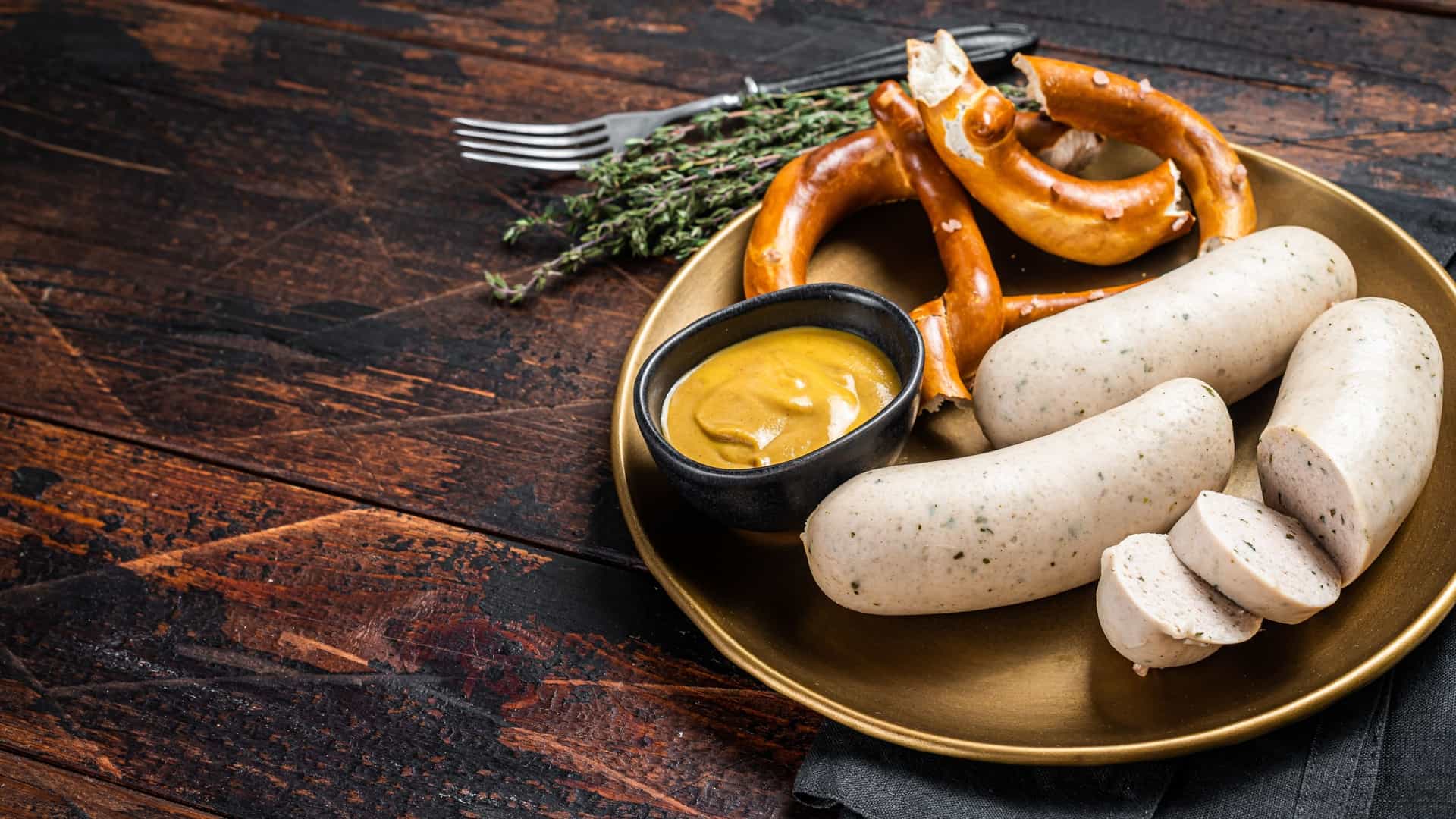 A top down view of a traditional Bavarian meal on a wooden table. The plate contains two Weisswurst, or white sausages, served with a pretzel and a small dish of sweet mustard.