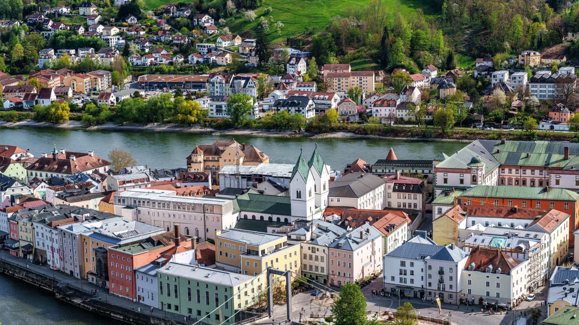 A high-angle view of the Dreiflüsseeck (Three Rivers Corner) in Passau, Germany, showing the dramatic confluence of the Danube, Inn, and Ilz rivers. The waters clearly maintain their distinct colors at the point of merging: the brown/blue water of the Danube is visibly separated from the lighter, often green-tinged water of the Inn. The confluence point is at the tip of the peninsula, and the waters flow together past a low-lying stone embankment with an older building complex (Veste Niederhaus) on the shore.