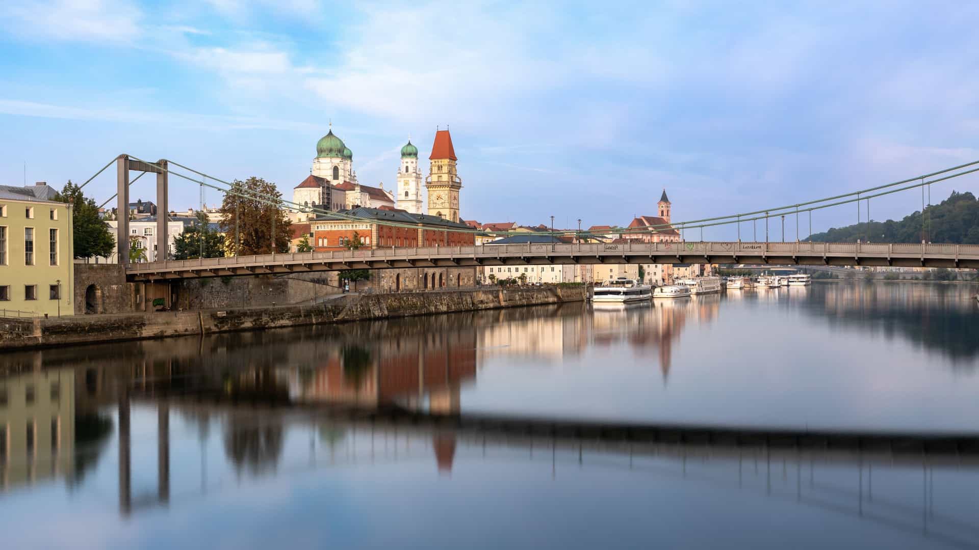 The Luitpold Bridge (Prinzregent-Luitpoldbrücke), an elegant metal suspension bridge, spans the Danube River in Passau, Germany. The bridge's supports and deck are reflected on the calm water, with the historic skyline of the Old Town visible in the background, dominated by the green onion domes of St. Stephen's Cathedral.