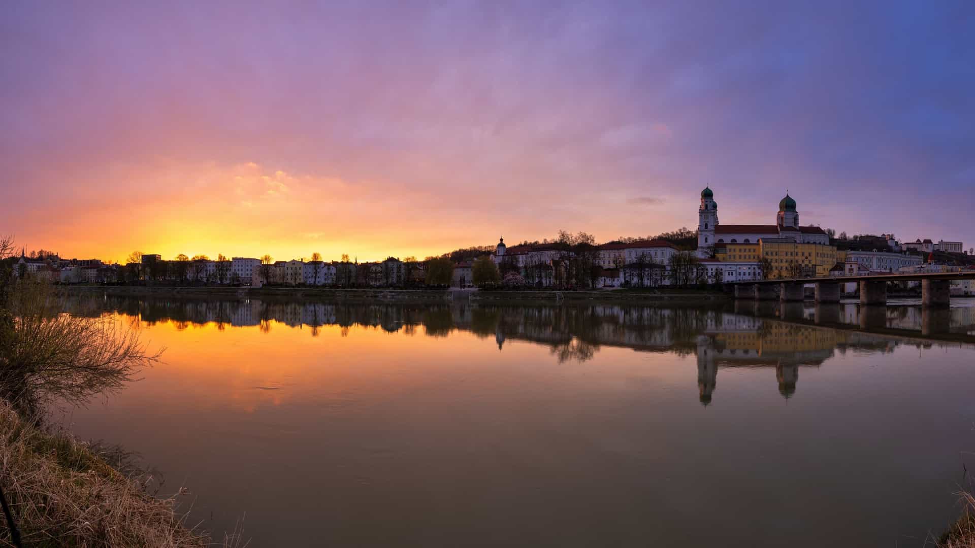 An incredible sunset view of Passau, Germany. The sky is a vibrant mix of orange, pink, and yellow hues, casting a warm glow over the city. The Danube river, with the historic old town and its Baroque architecture, reflects the colors of the sky.