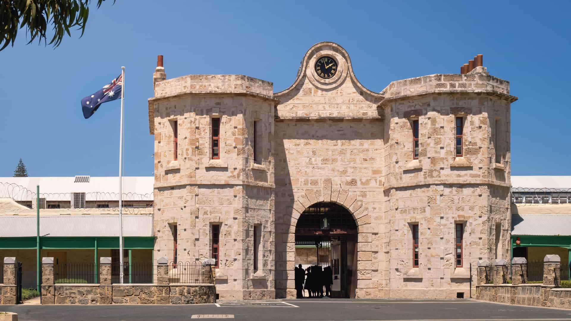 An image of the entrance to Fremantle Prison in Fremantle, Western Australia. The entrance is a large stone gatehouse with two rounded towers and a clock at the top, leading into the former prison, which is a World Heritage Site.