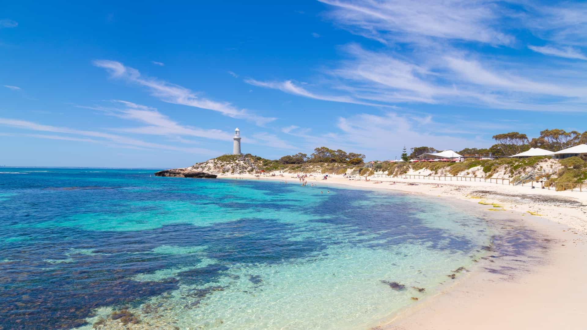 A scenic view of Pinky Beach on Rottnest Island, Western Australia. The image shows the white sand and clear turquoise water, with the Bathurst Lighthouse visible in the background. The beach is known for its calm waters and is a popular spot for swimming and watching the sunset.
