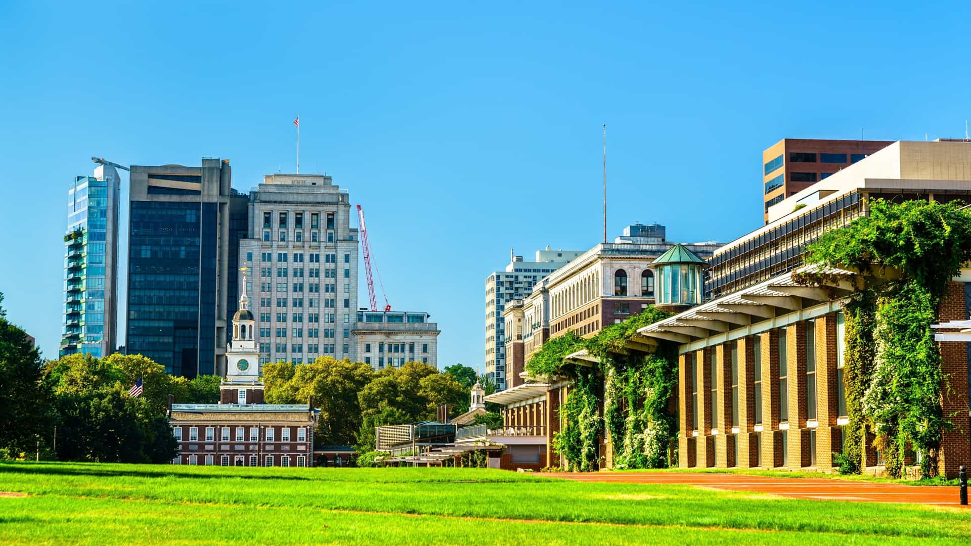 Independence Hall, a red brick building with a bell tower and clock, is the central focus of Independence National Historical Park in Philadelphia. It stands on a green lawn, surrounded by trees under a clear blue sky.