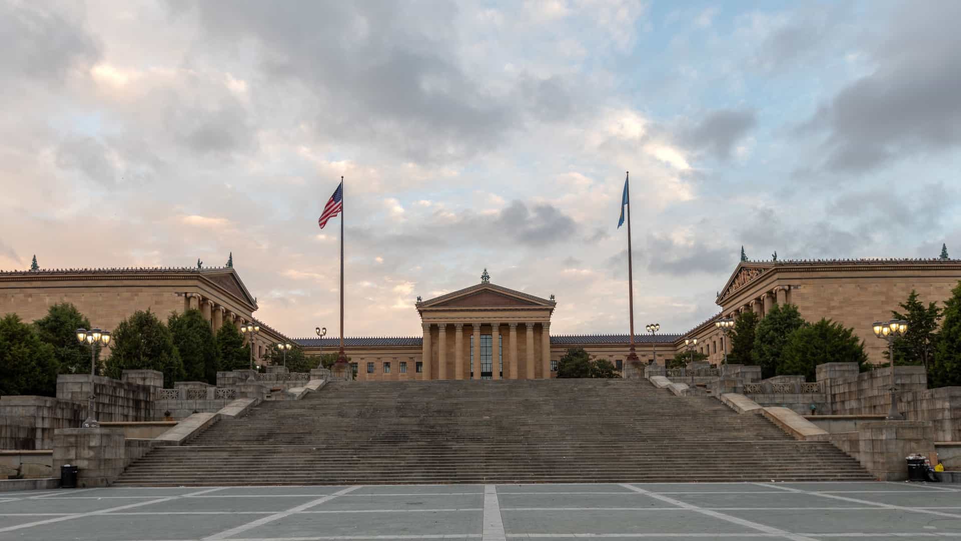 A wide angle view of the Philadelphia Museum of Art, a prominent Georgian style building with a classical facade and large columns, seen from the bottom of the "Rocky Steps." The museum is situated at the top of a wide, grand set of stairs, and a fountain and trees are visible in the foreground, with the city skyline in the distance.