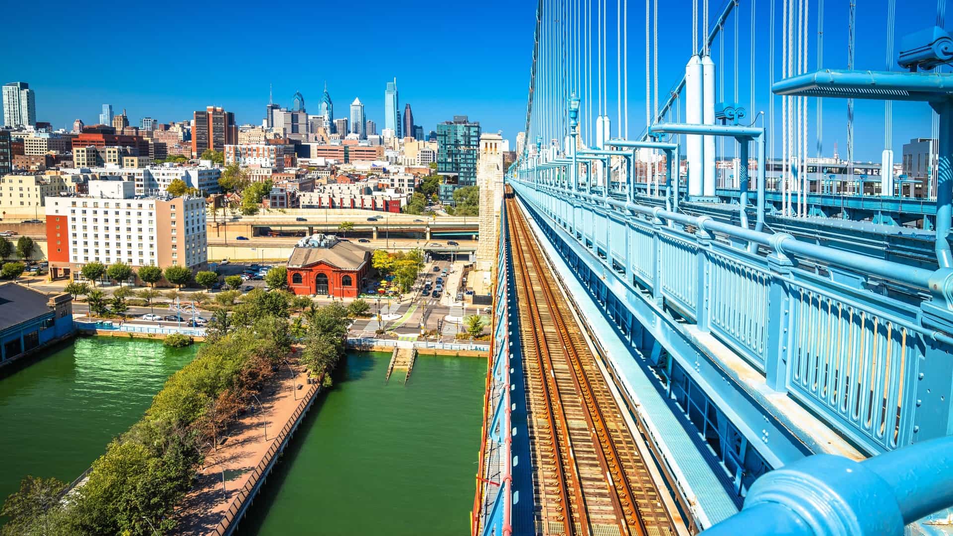 A wide angle view of the Philadelphia skyline from the Benjamin Franklin Bridge, with the Delaware River waterfront in the foreground. The city's skyscrapers rise above the river, and the bridge's suspension cables are a prominent feature of the scene.