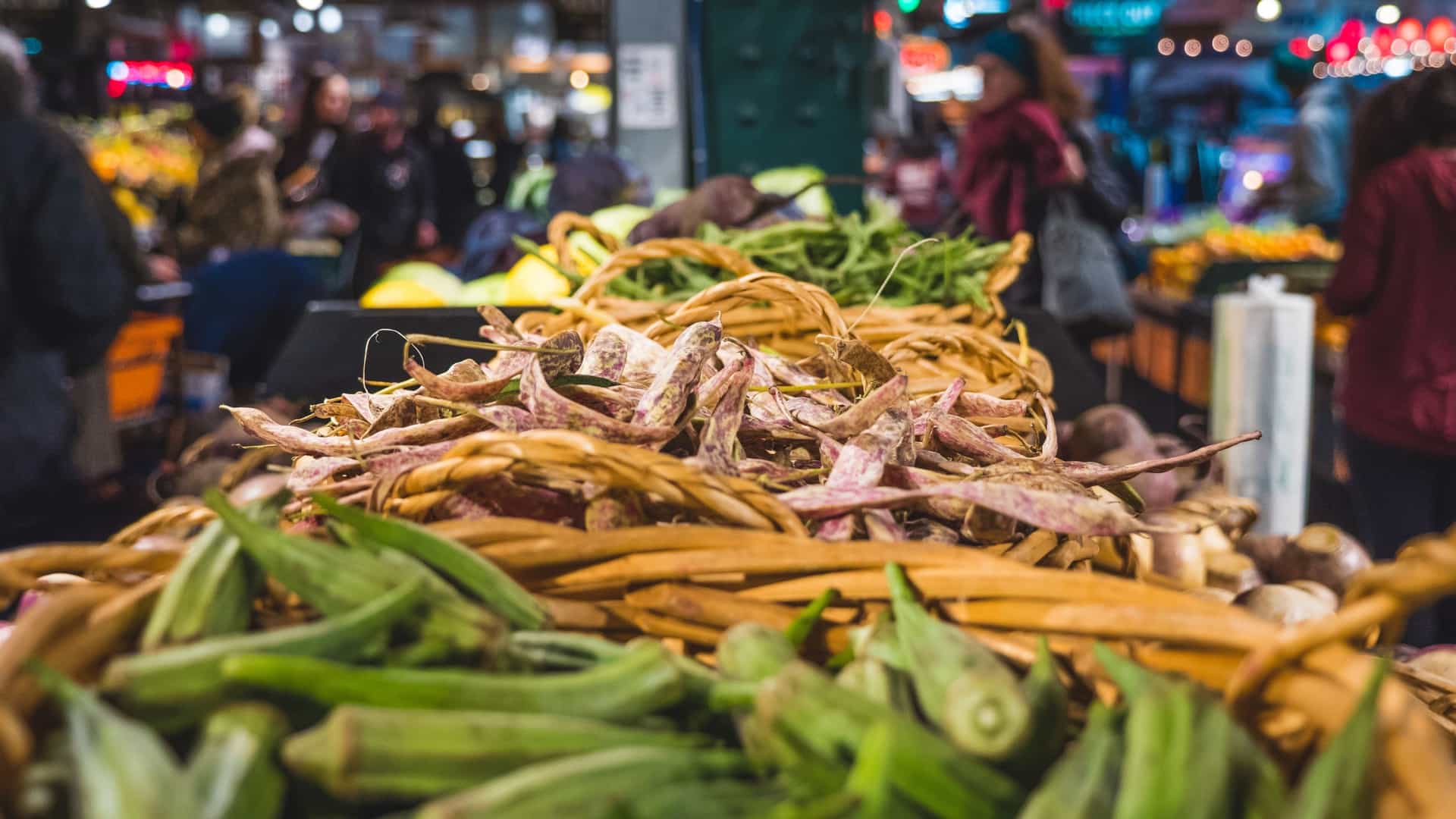 A view inside the historic Reading Terminal Market in Philadelphia, showing the wide central aisle of the bustling indoor public market. The photo captures several vendor stalls displaying fresh groceries, produce, and specialty foods under the market's distinctive high, arched ceiling and historic architecture, with many people walking and shopping.