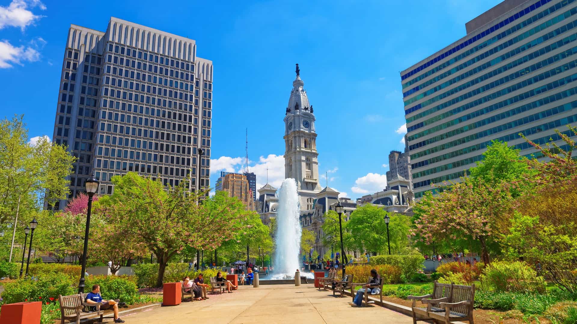 A view of Love Park in Philadelphia, featuring its central fountain with water jets. In the background, the iconic Philadelphia City Hall building is visible, with its detailed architecture and clock tower. People are seen relaxing in the park and near the fountain.