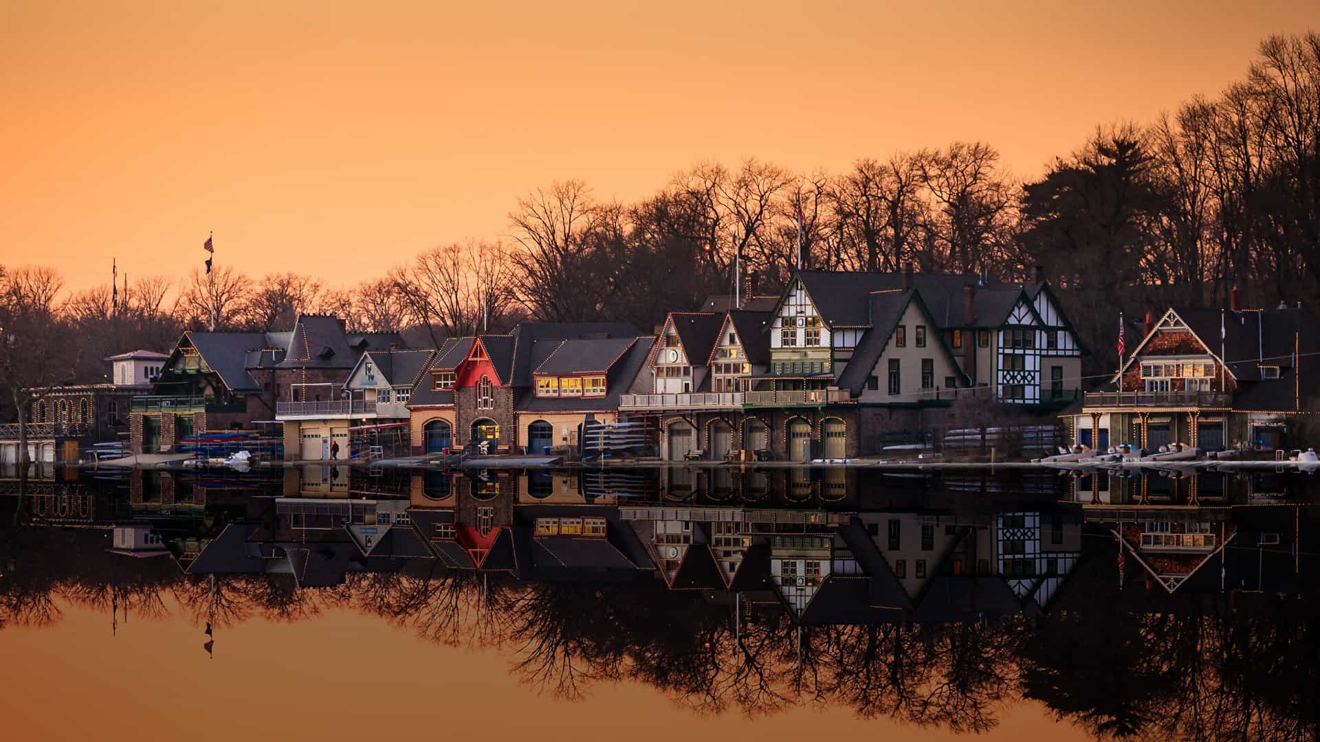 Boathouse Row at sunset in Philadelphia, with the historic 19th century boathouses illuminated by lights. The colorful buildings are reflected in the tranquil water of the Schuylkill River, and a vibrant sky with a mix of orange and purple hues is visible in the background.