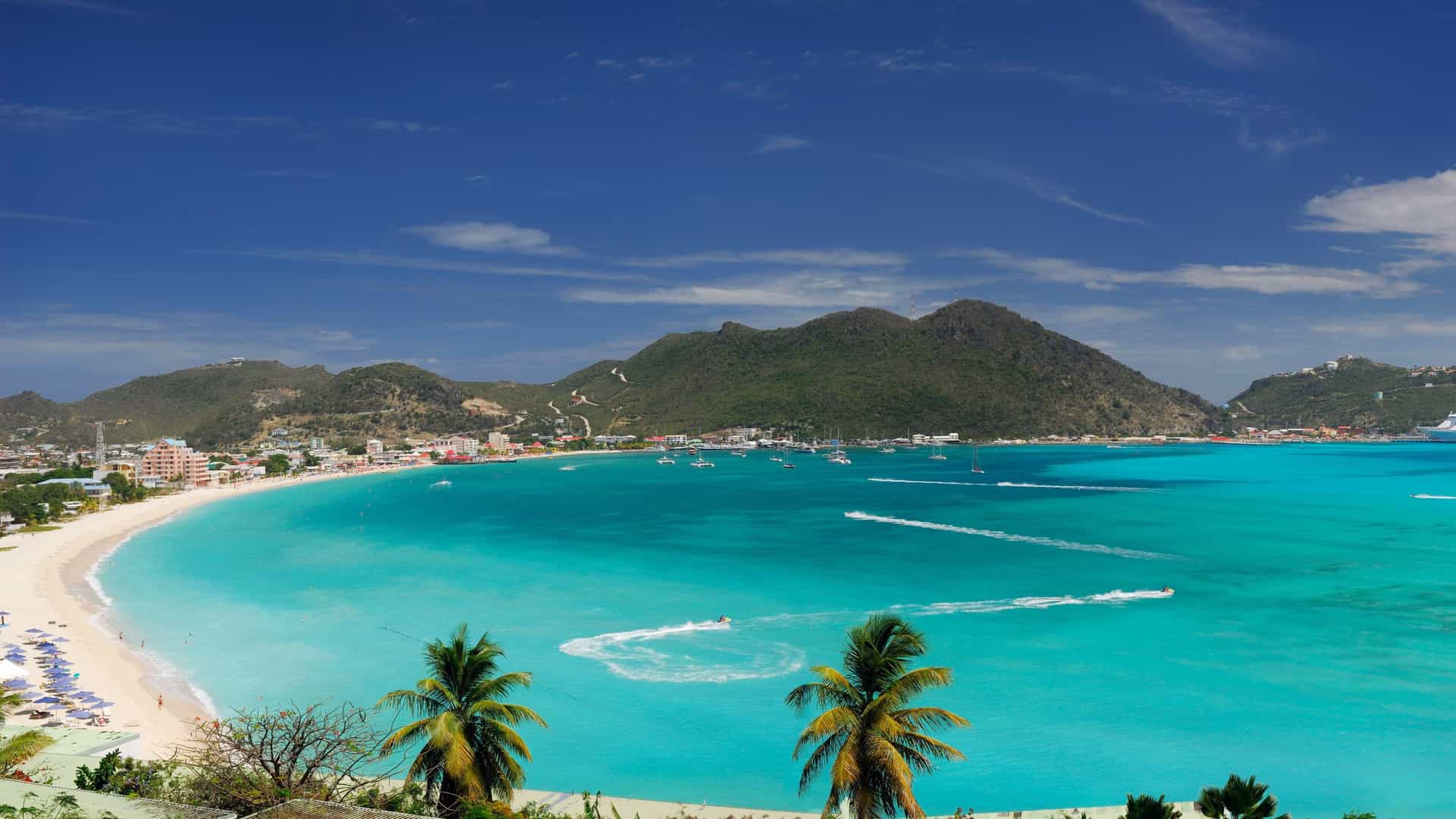 A panoramic view of Great Bay Beach in Philipsburg, St. Maarten. The image shows a wide expanse of white sand beach dotted with sun loungers and colorful umbrellas. The turquoise water of the Caribbean Sea is calm, and a wooden dock extends from the shore. Behind the beach, a row of bright, colorful buildings lines a boardwalk, and lush green mountains rise in the background. Several large cruise ships are anchored in the distance.
