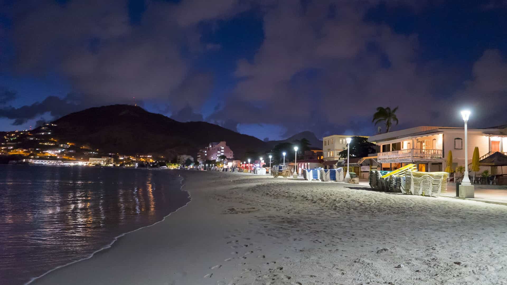 A nighttime view of the Philipsburg Boardwalk and Great Bay Beach in St. Maarten. The boardwalk is illuminated by numerous streetlights and the lights from the restaurants and shops lining the street. The lights reflect on the wet surface of the wooden boardwalk and the calm, dark water of the bay. The scene is quiet, suggesting the late hour.